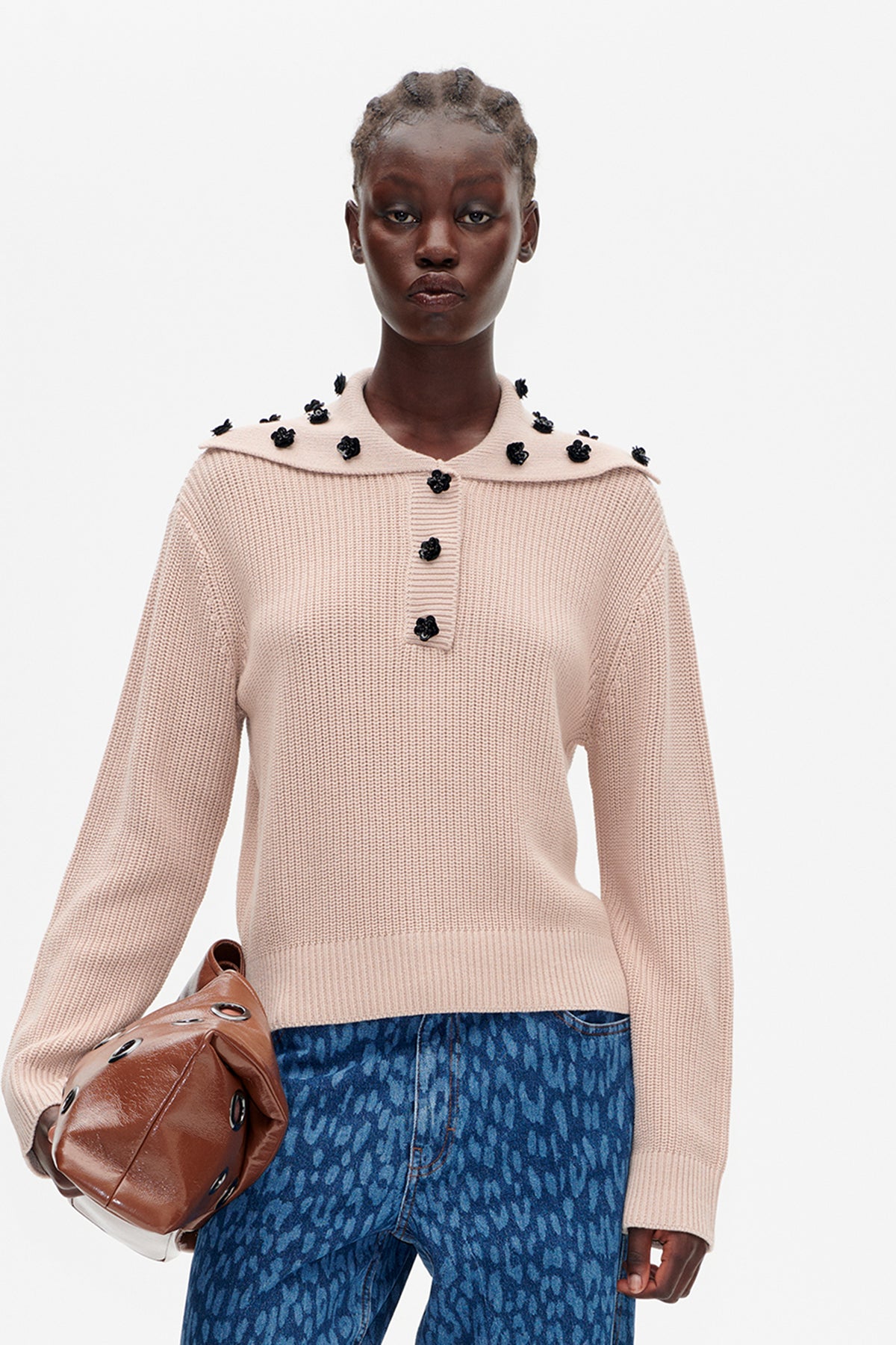 Model wearing a pale pink textured knit sweater with a wide collar and black floral embellishments, paired with patterned blue jeans.