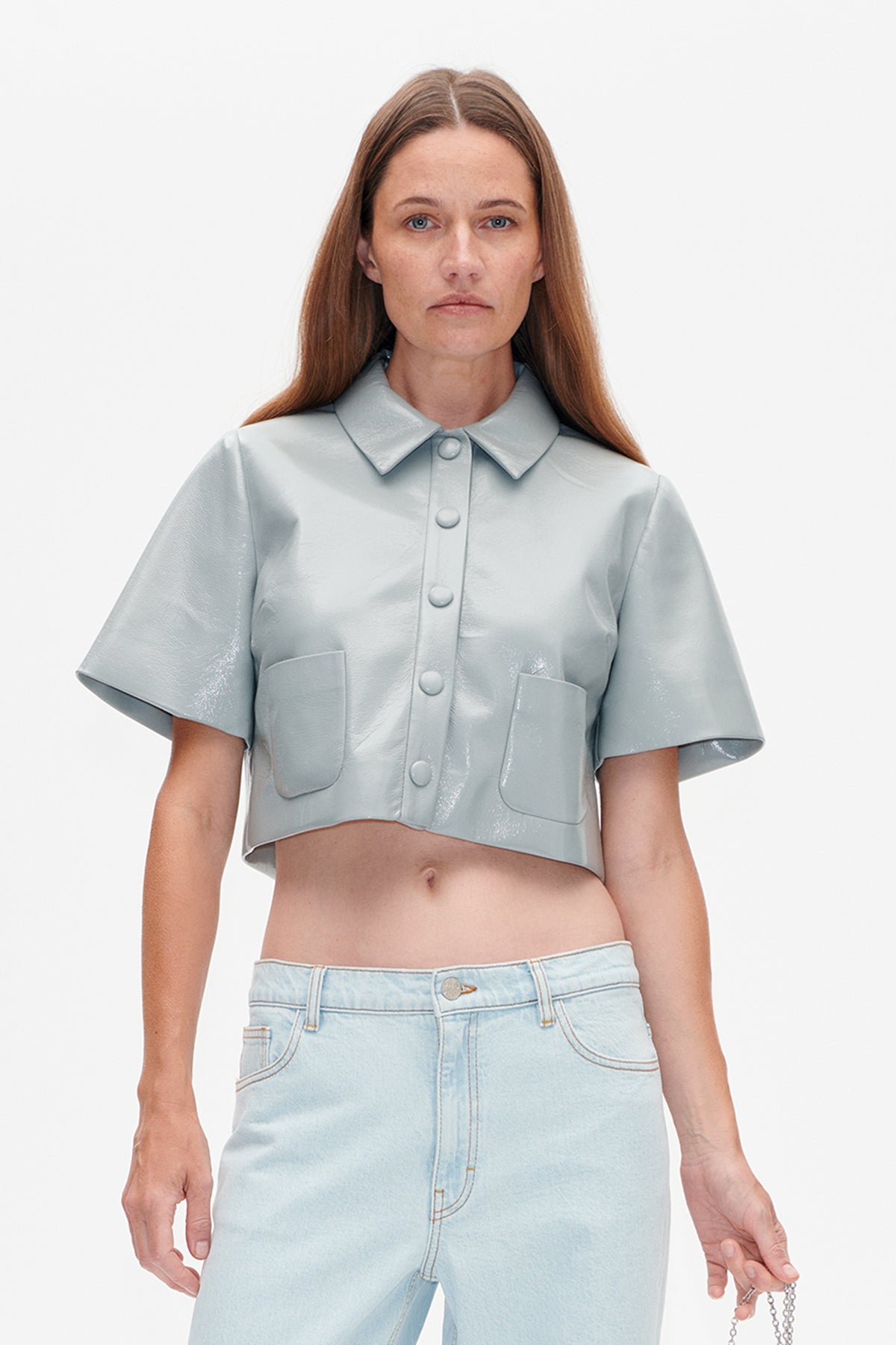 Front view of a pale blue glossy cropped shirt with short sleeves, pointed collar, matching buttons and two patch pockets, styled with light-wash jeans.