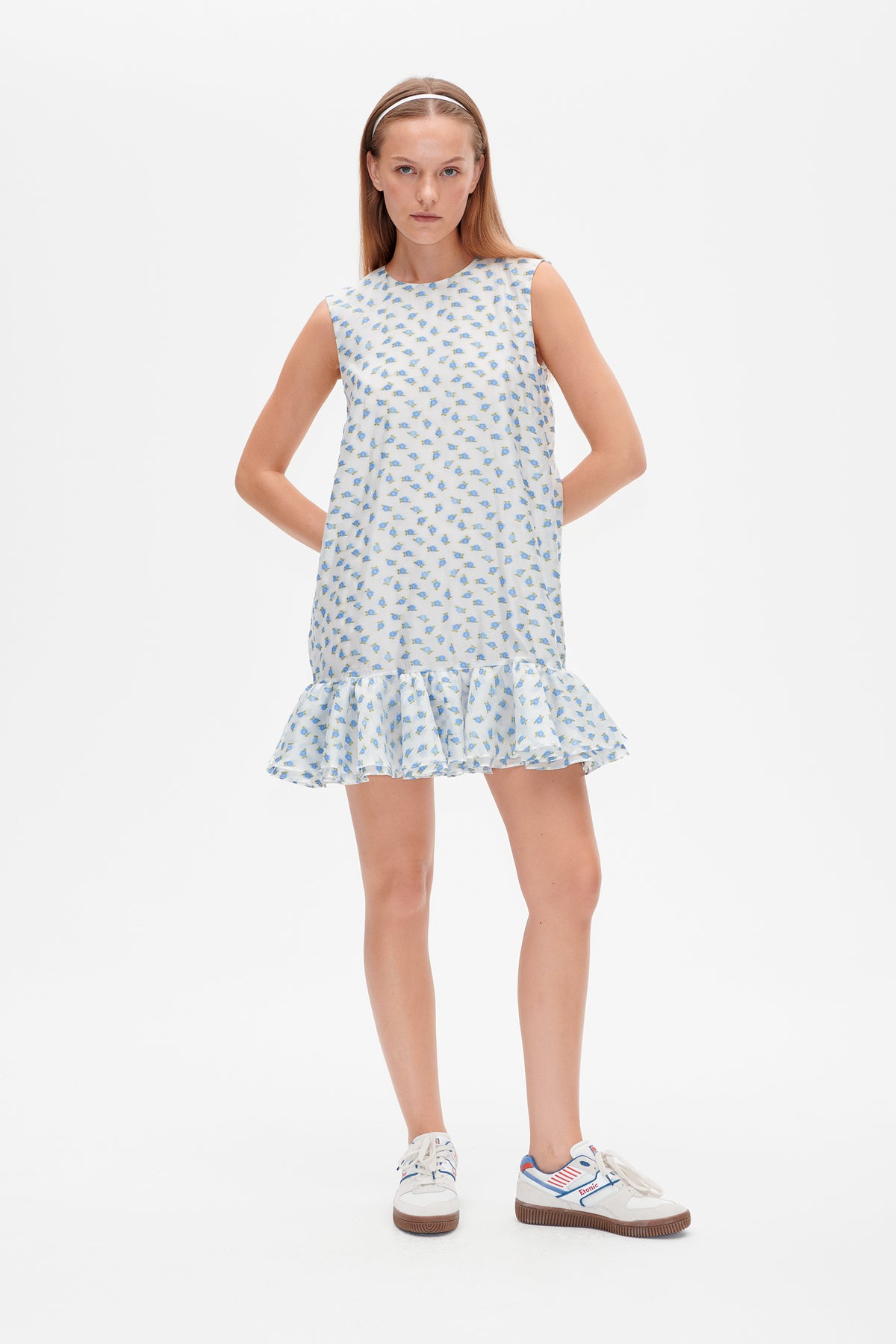 Model wearing a sleeveless white mini dress with a blue and green floral print and a ruffled hem, shown from the front.