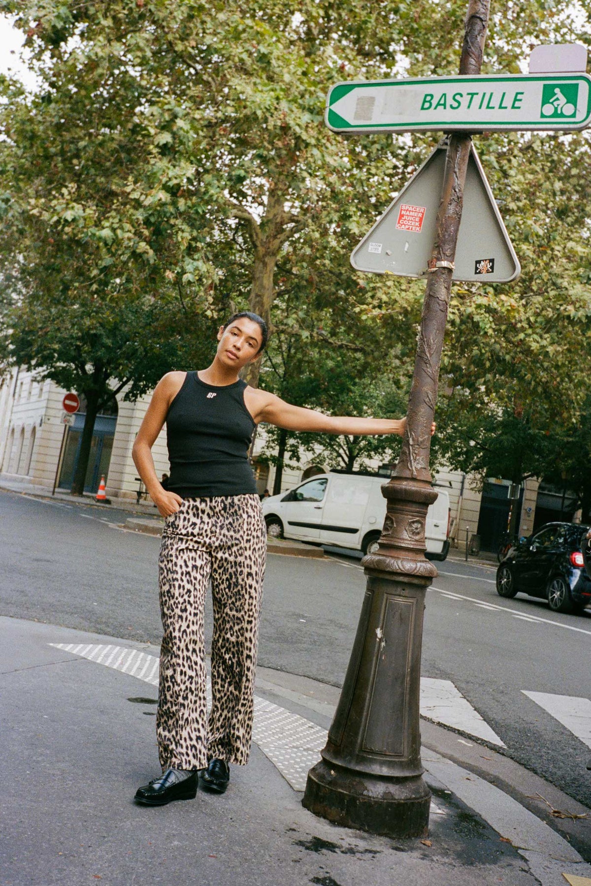 Een model poseert bij een Bastille-bord in een donkere mouwloze top en een losse beige broek met zwarte luipaardprint.