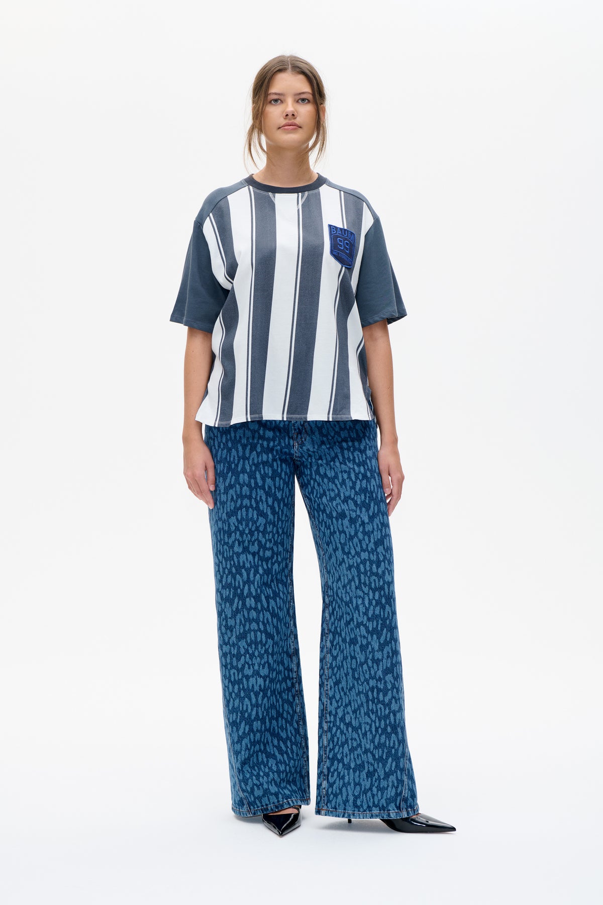 Vorderansicht eines Models, das ein kurzärmliges, blau-weiß vertikal gestreiftes T-Shirt mit Brustpatch und eine gemusterte blaue Wide-Leg-Jeans trägt.