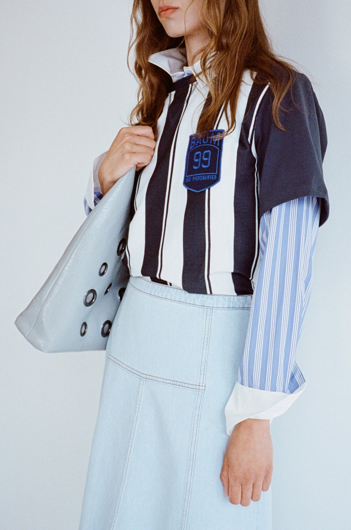 Beschnittener Kampagnenausschnitt eines Models, das ein ärmelloses, marineblau-weiß gestreiftes Top über einer blau gestreiften Bluse trägt, dazu einen hellen denim-Rock und eine graue Tote-Bag.