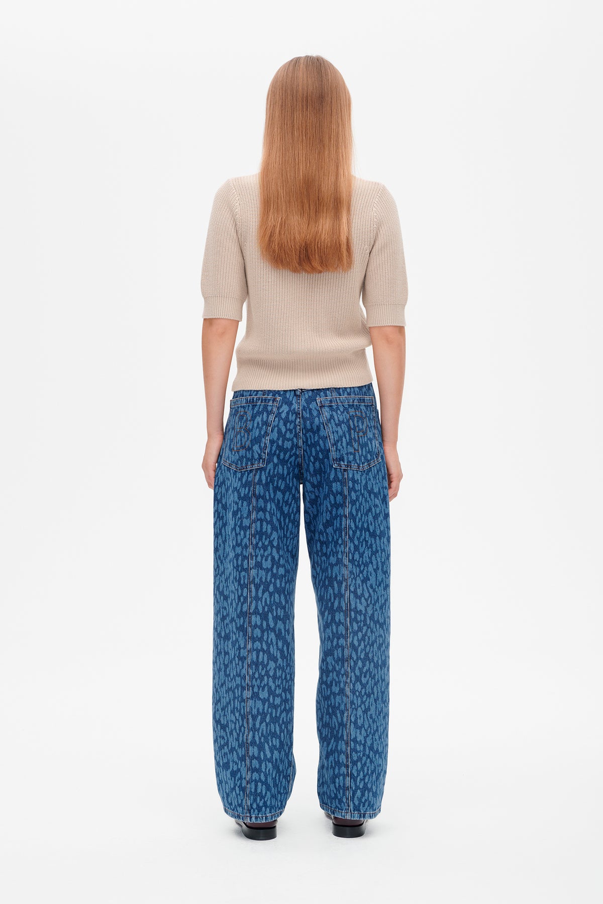 Rückenansicht eines Models, das ein beiges kurzärmeliges Stricktop und blaue Jeans mit weitem Bein, Allover‑Muster und Gesäßtaschen trägt.