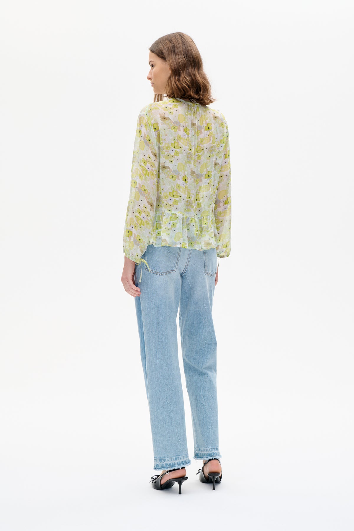 Model van achteren gezien draagt een lichte, doorschijnende blouse met een geelgroene bloemenprint, lange mouwen en een geplooide peplumzoom, gecombineerd met lichtblauwe jeans.