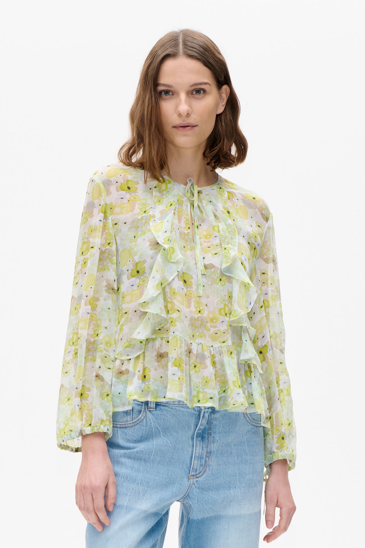 Front view of a pale blouse with a yellow and green abstract floral print, tie neckline, cascading ruffles and gathered three-quarter sleeves.