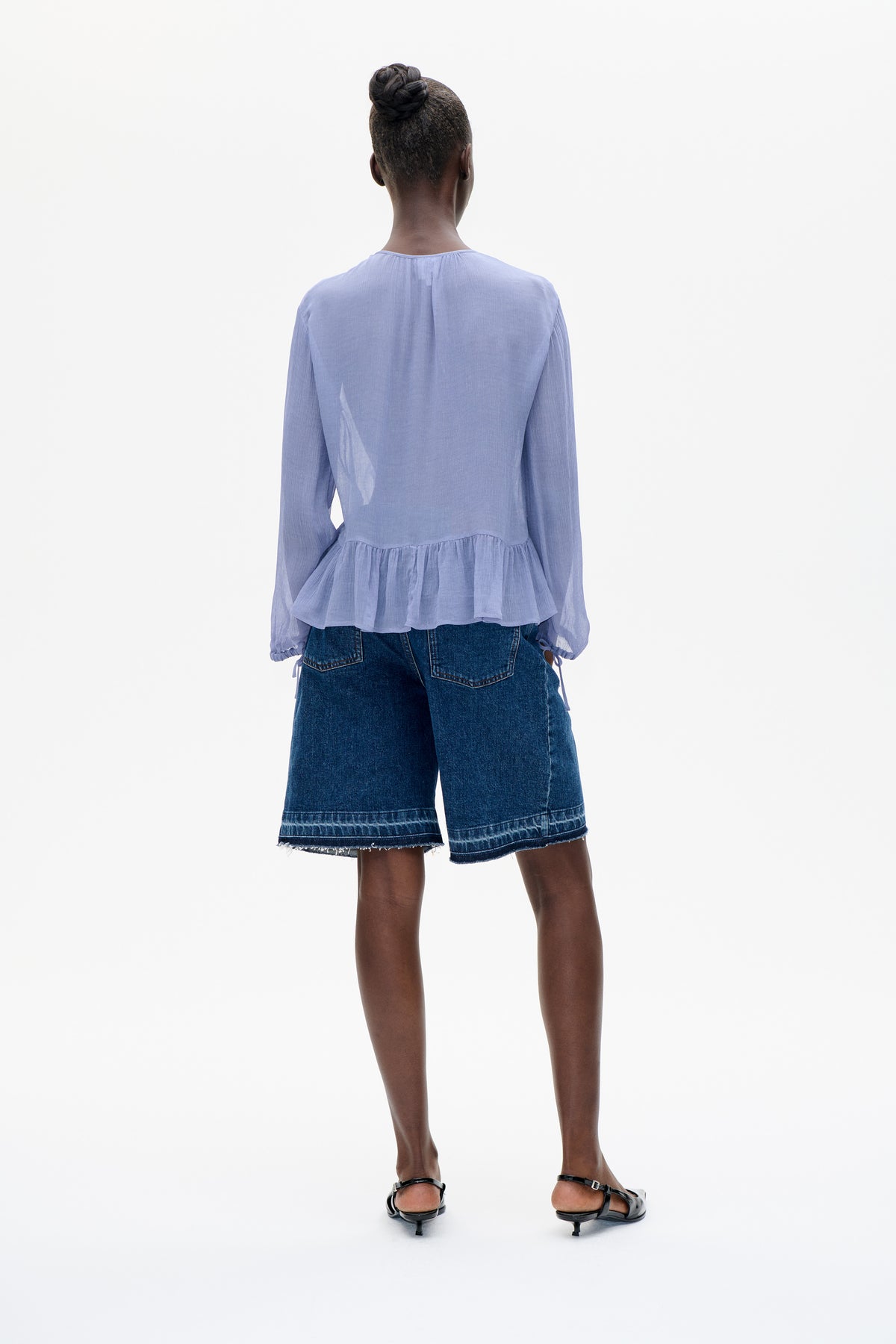 Achteraanzicht van een licht lavendelkleurige blouse met lange mouwen en een gerimpelde peplumzoom, gecombineerd met donkere denim shorts.