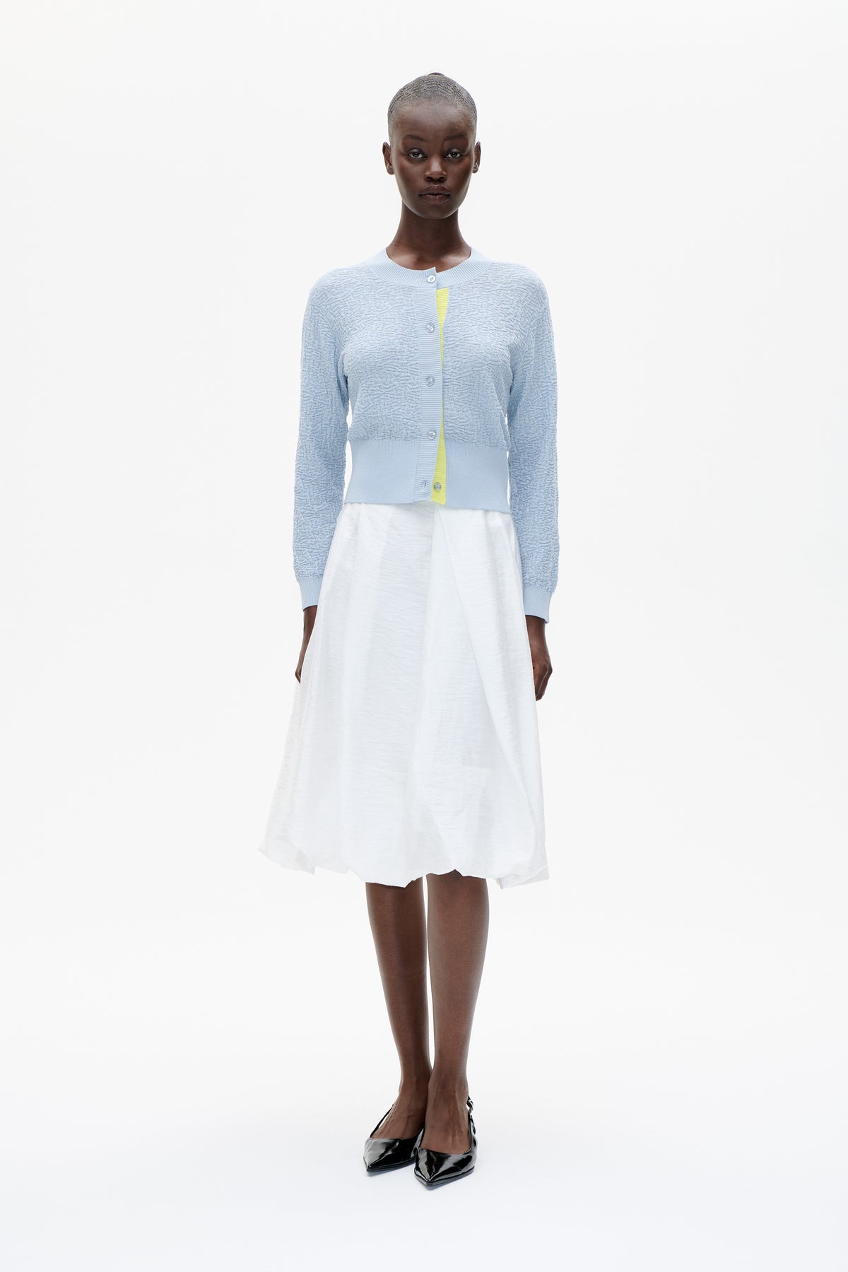 Front view of a model wearing a pale blue textured button-front cardigan over a white dress, styled with black pointed shoes.
