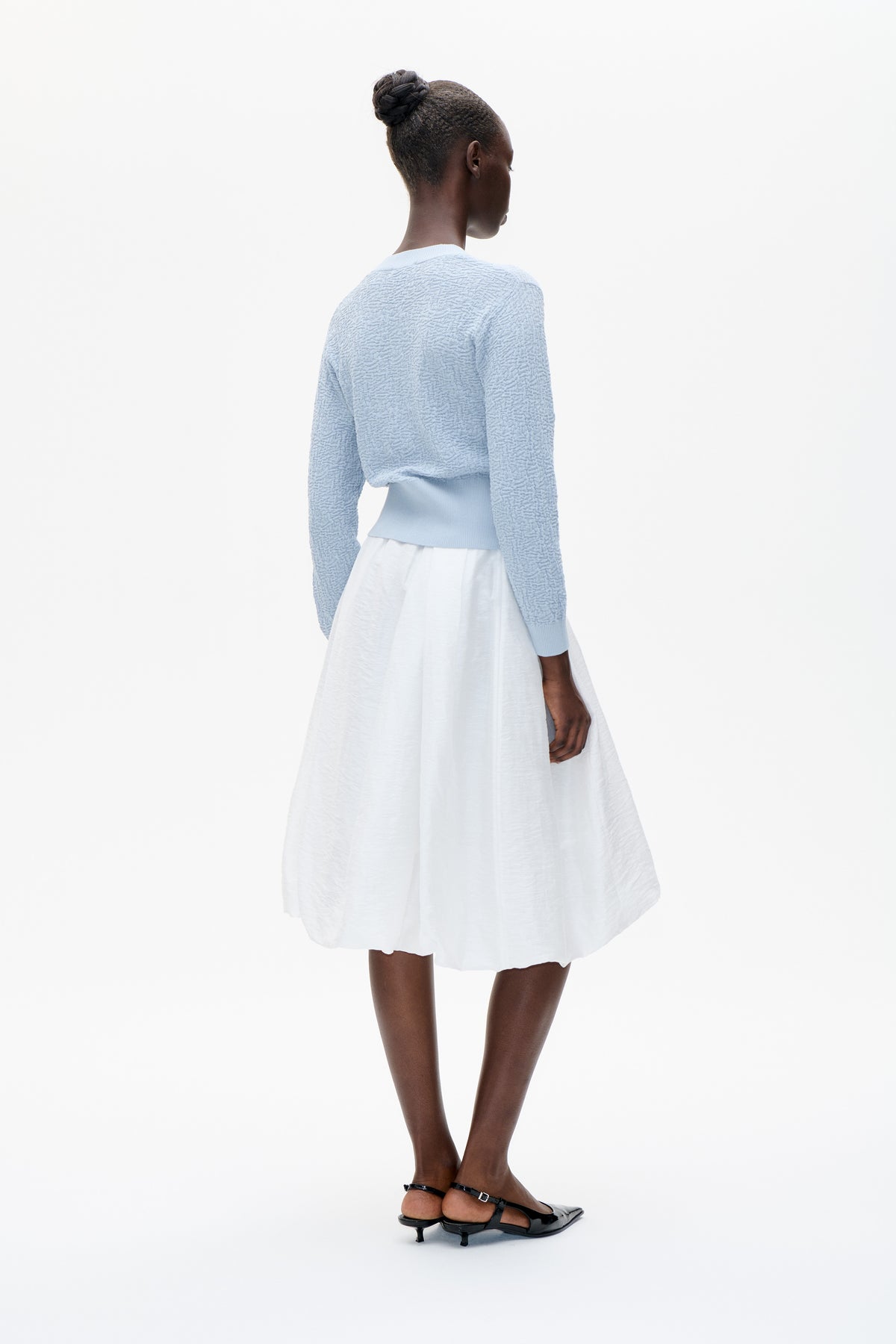 Back view of a model wearing a pale blue textured knit sweater with ribbed cuffs and hem, paired with a flowing white skirt.