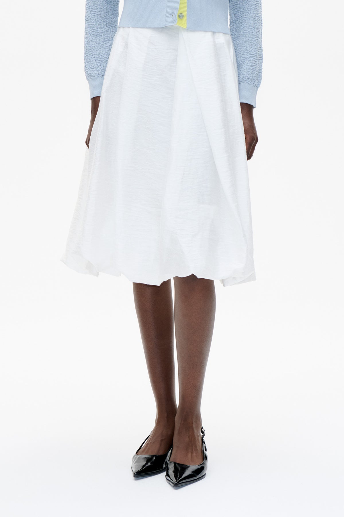 Front detail of a white flared skirt with subtle pleats, paired with a pale blue textured cardigan featuring a yellow button placket.
