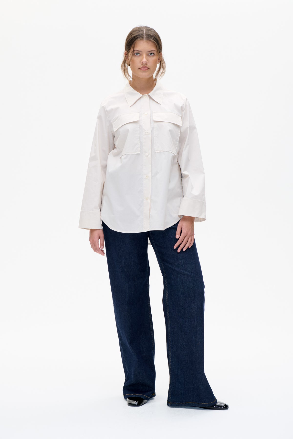 Front view of a model wearing an off-white button-front shirt with a pointed collar and four large flap pockets, paired with dark wide-leg jeans.
