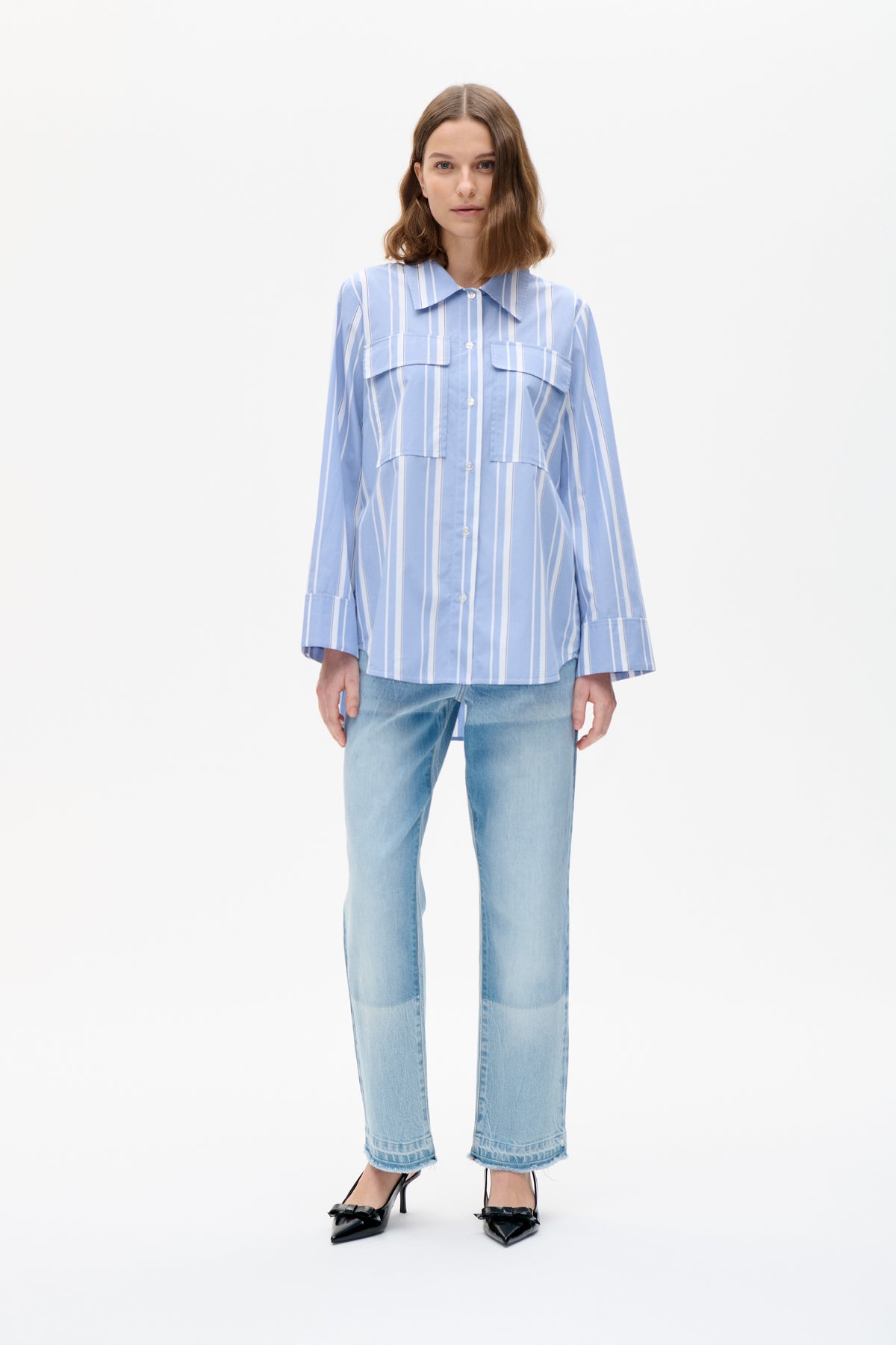 Model trägt ein blau-weiß vertikal gestreiftes, geknöpftes Hemd mit spitzem Kragen und Brusttaschen mit Klappen, kombiniert mit hellblauen Jeans.
