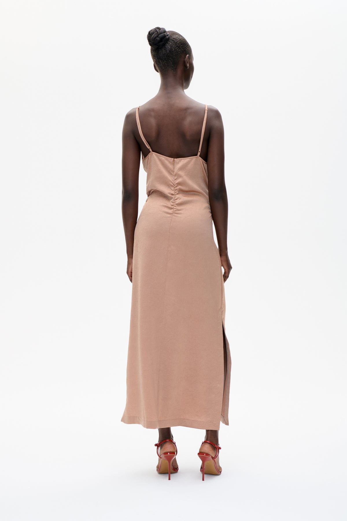 Back view of a model wearing a blush pink slip dress with thin adjustable straps, a ruched back seam and side slit.