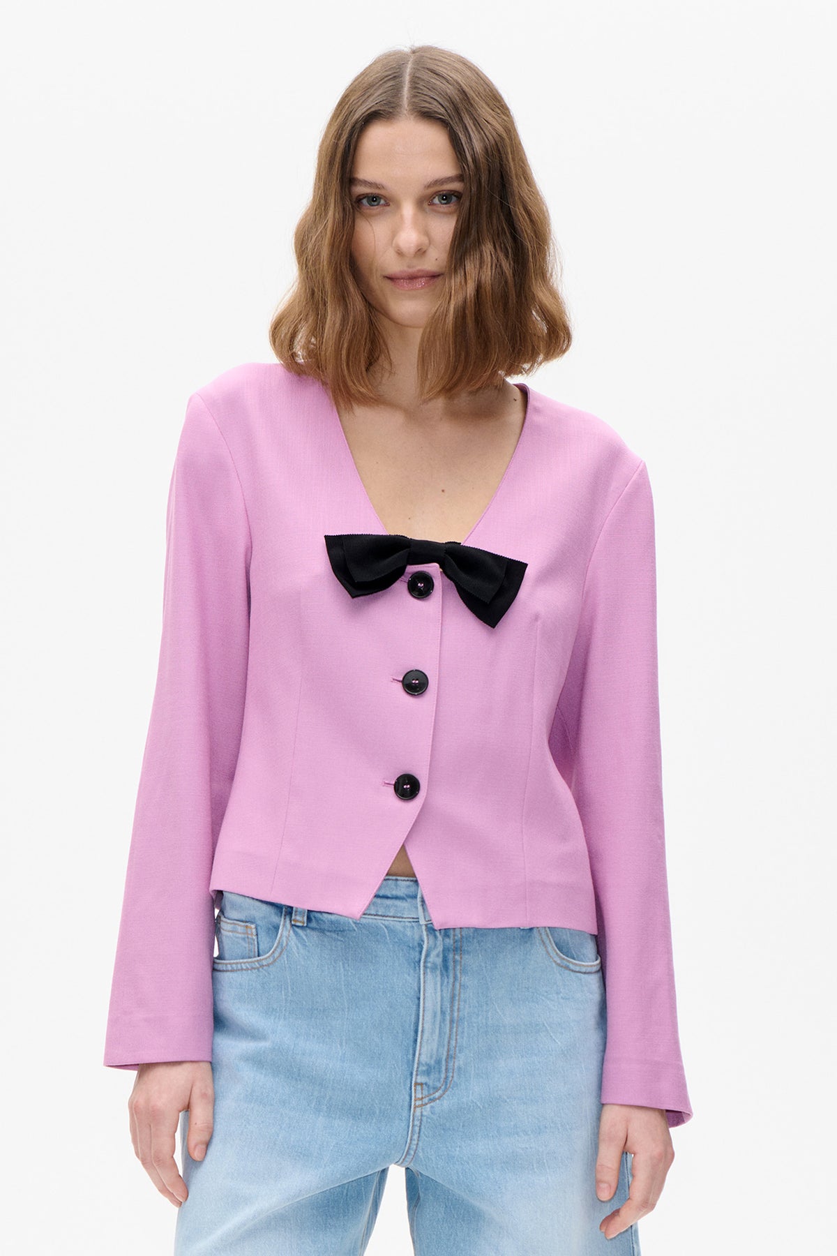 Front detail of a pink V-neck jacket with black buttons and a large black bow, styled with light-wash jeans.