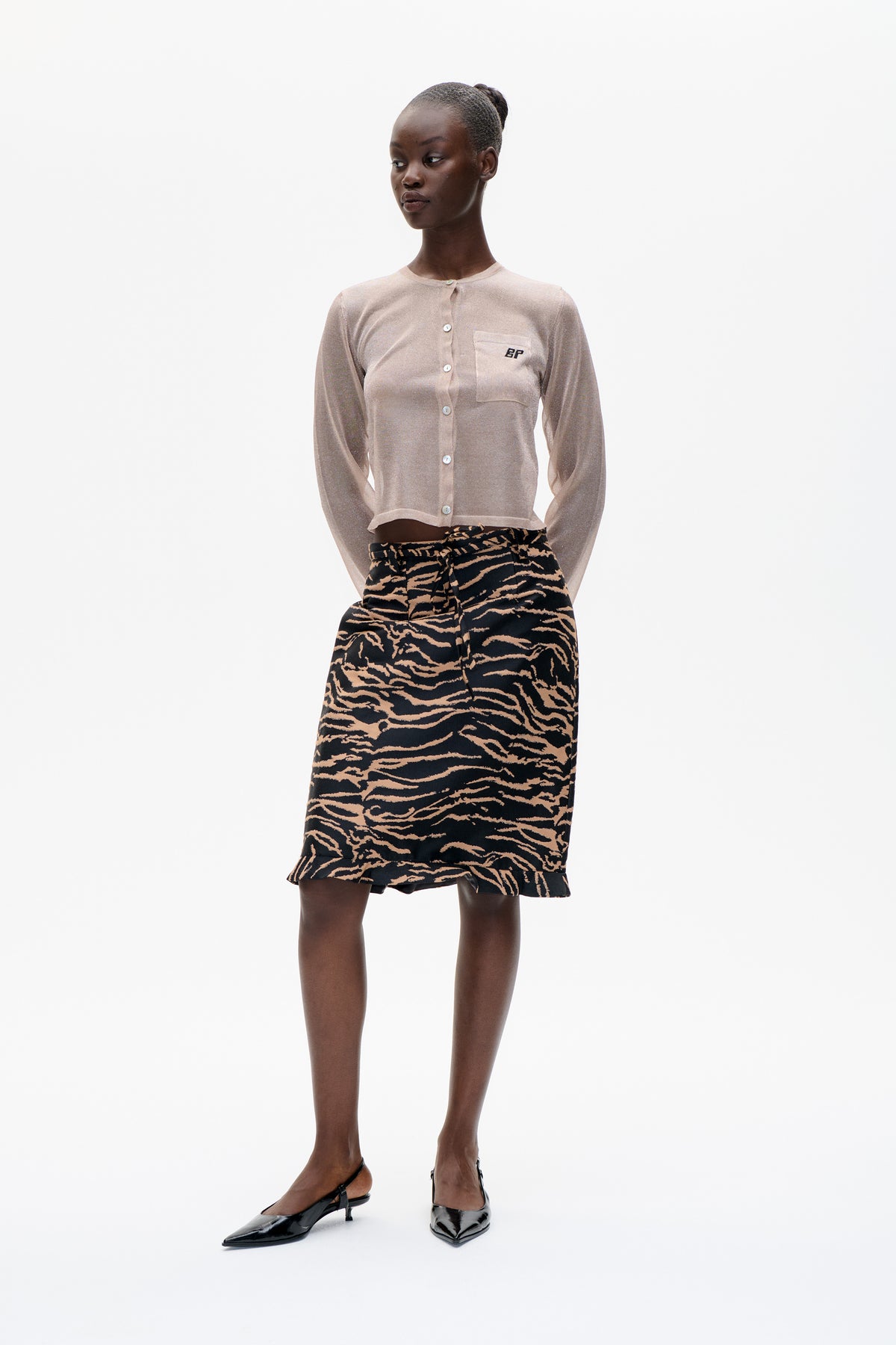 Model wearing a pale blush button-front cardigan with a chest pocket and a black skirt patterned with tan wavy stripes.