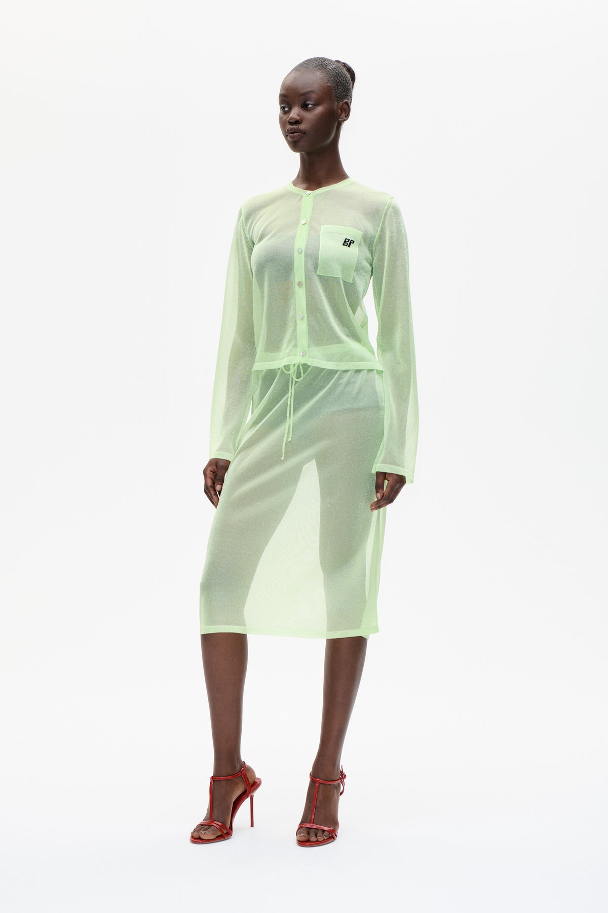 Full-length studio portrait wearing a pale lime sheer button-front cardigan with a BP chest pocket, matching sheer bottoms and red strappy heels.