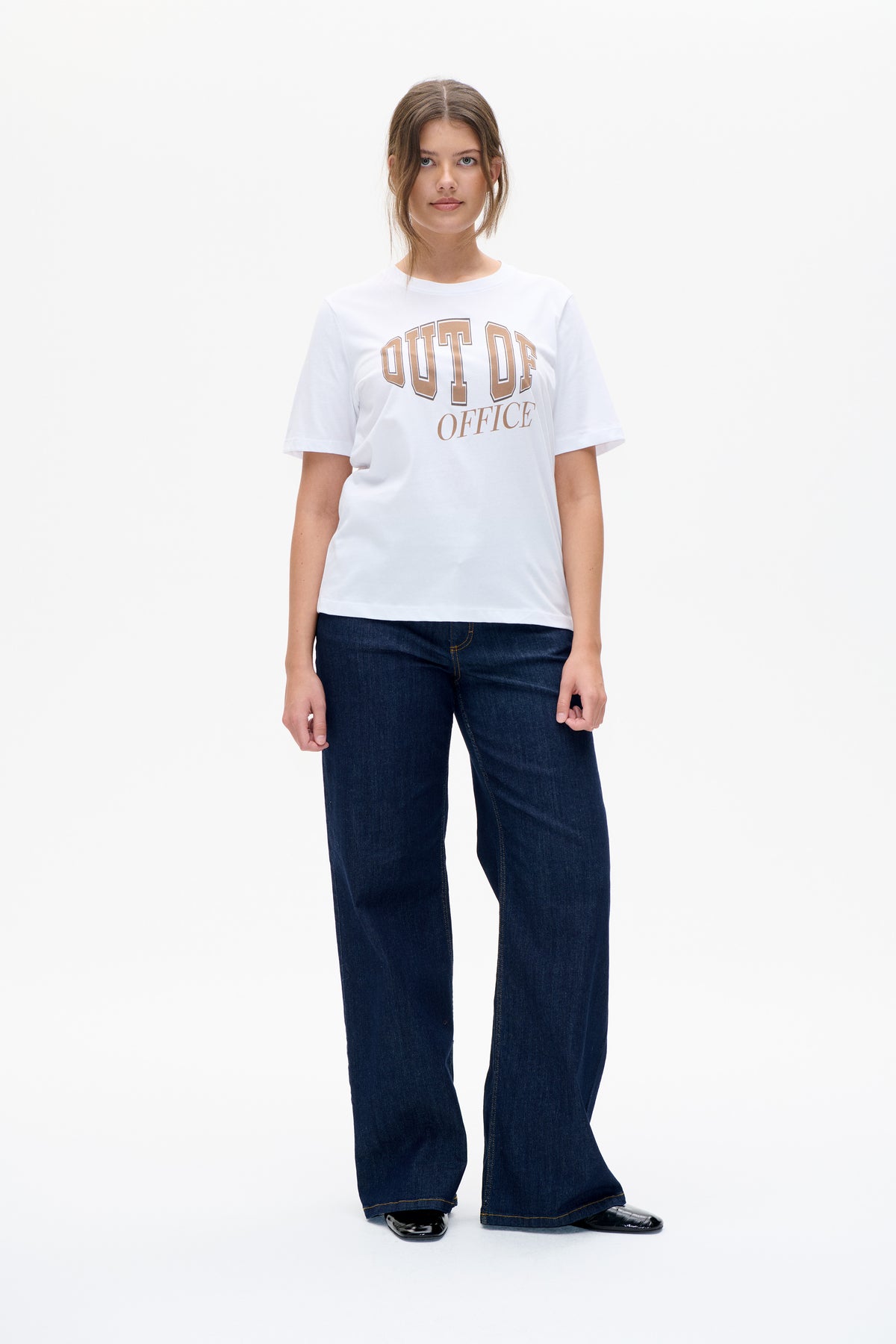 Front view of a model wearing a white short-sleeve T-shirt with tan “OUT OF OFFICE” lettering and dark wide-leg jeans.