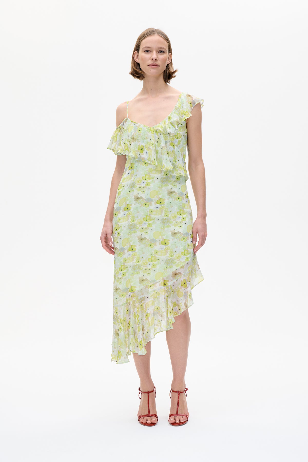 Model wearing an asymmetrical ruffled dress with thin straps and a pale yellow-green floral print, shown from the front.