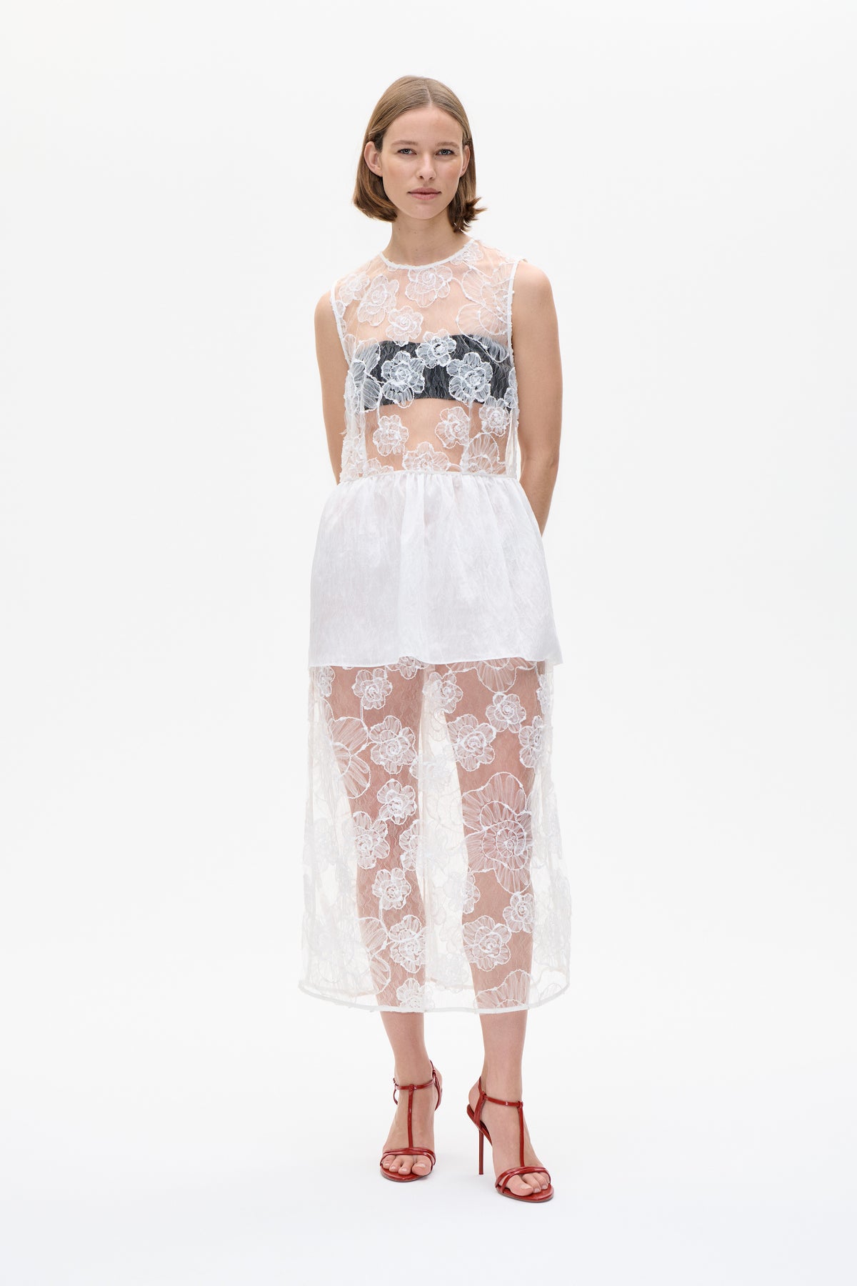 Front view of a white sleeveless dress with a sheer floral lace bodice, black bandeau underneath and layered skirt.