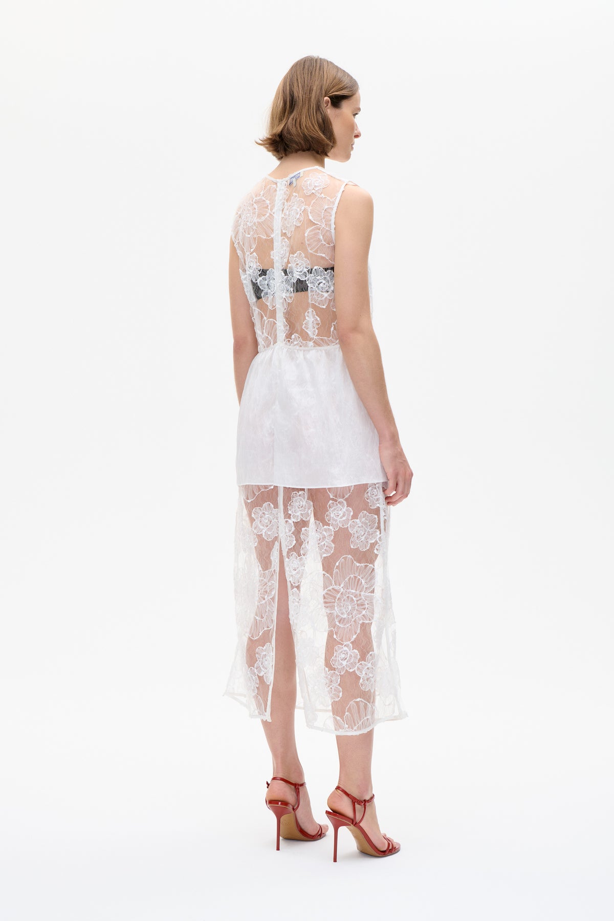 Model seen from behind in a white sheer floral lace dress with a short opaque underlayer and high slit.