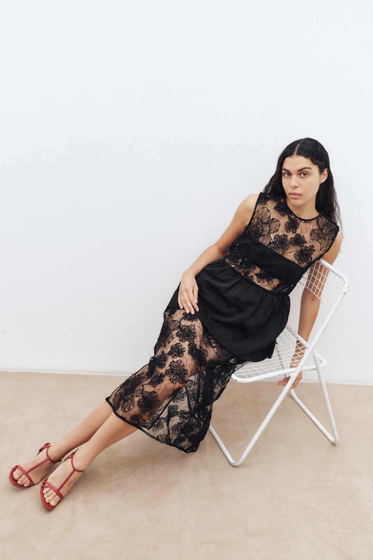 Model seated in a white wire chair wearing a black dress with a sheer floral lace bodice and skirt panels.