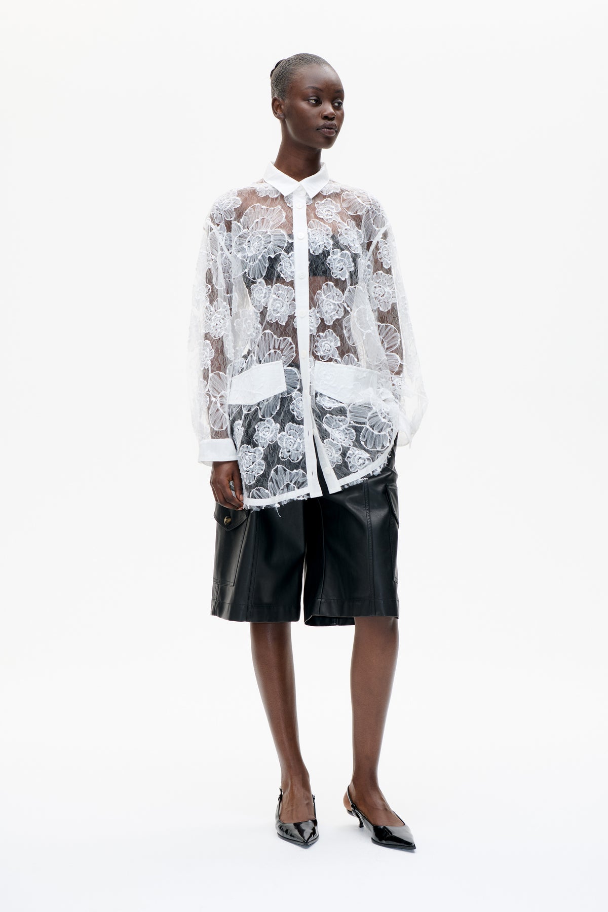 Model trägt transparente weiße Bluse aus floraler Spitze mit Knopfleiste und spitzem Kragen, dazu schwarze knielange Shorts vor weißem Hintergrund