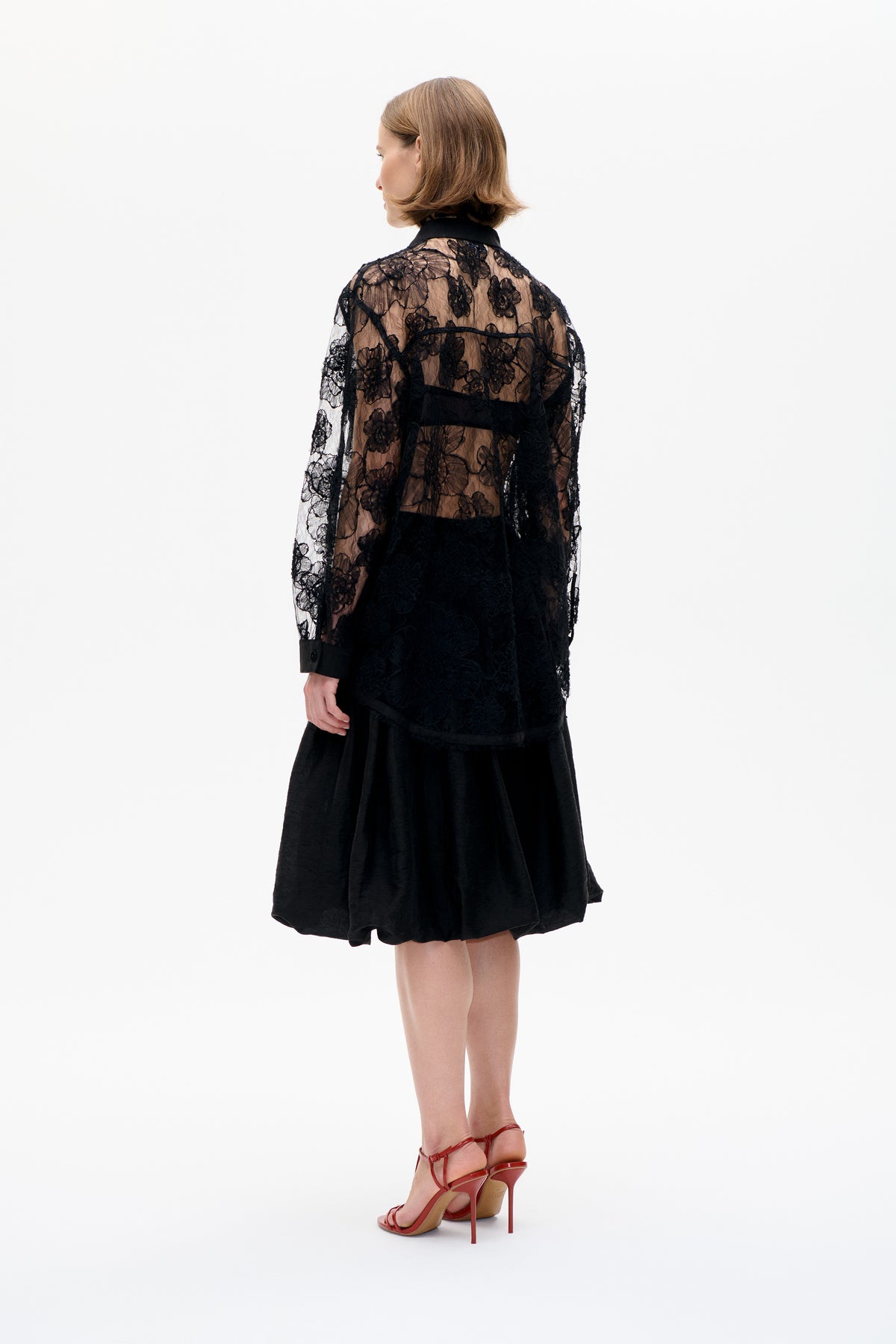 Model shown from behind in a sheer black floral lace blouse with a solid collar, cuffs and lower panel, paired with a black skirt.