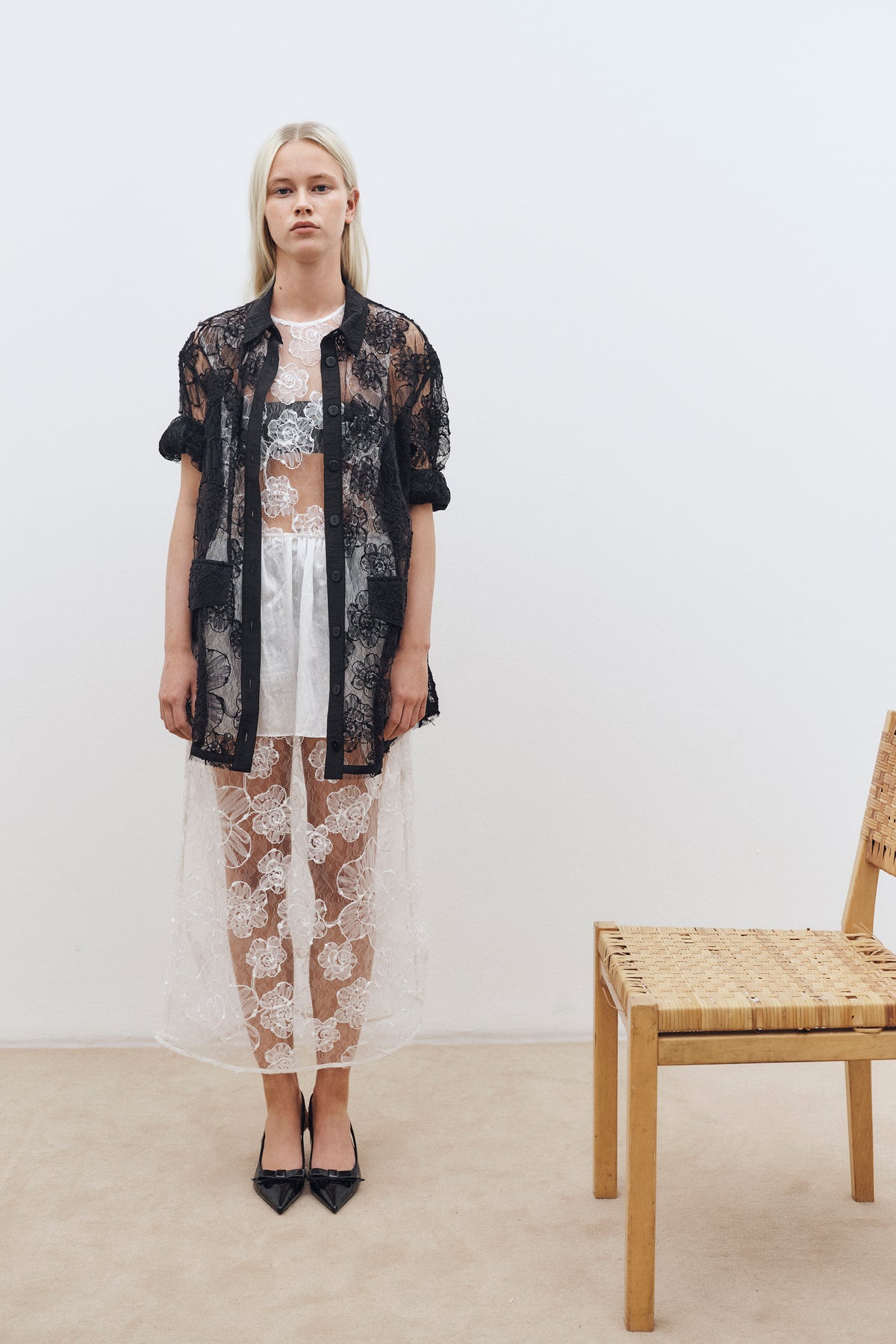 Model stands beside a woven chair wearing a sheer black floral lace button-front shirt over a white floral lace dress.