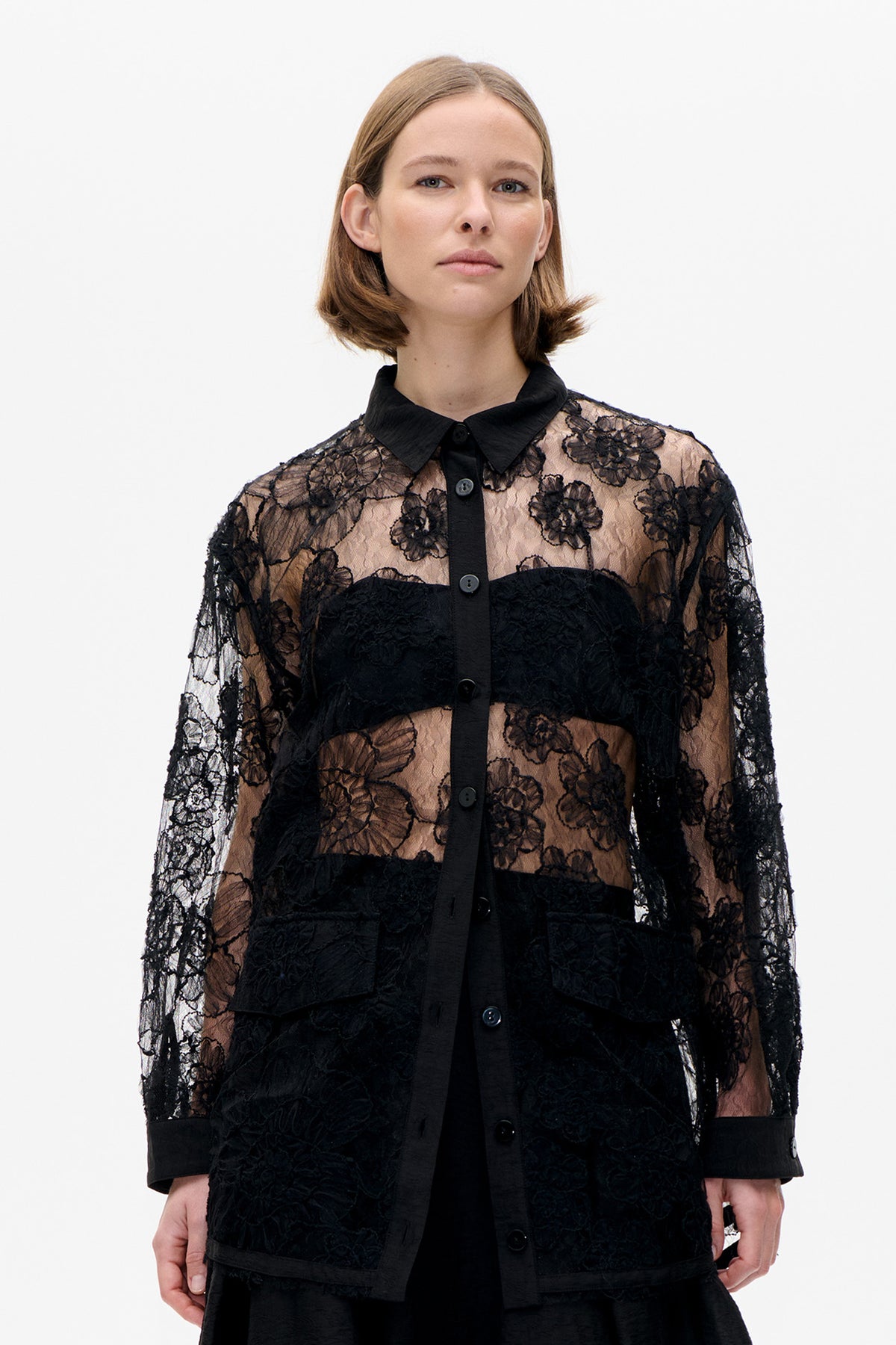 Front detail of a black button-front blouse with sheer floral lace sleeves and panels, opaque black bands, pointed collar and wide cuffs.