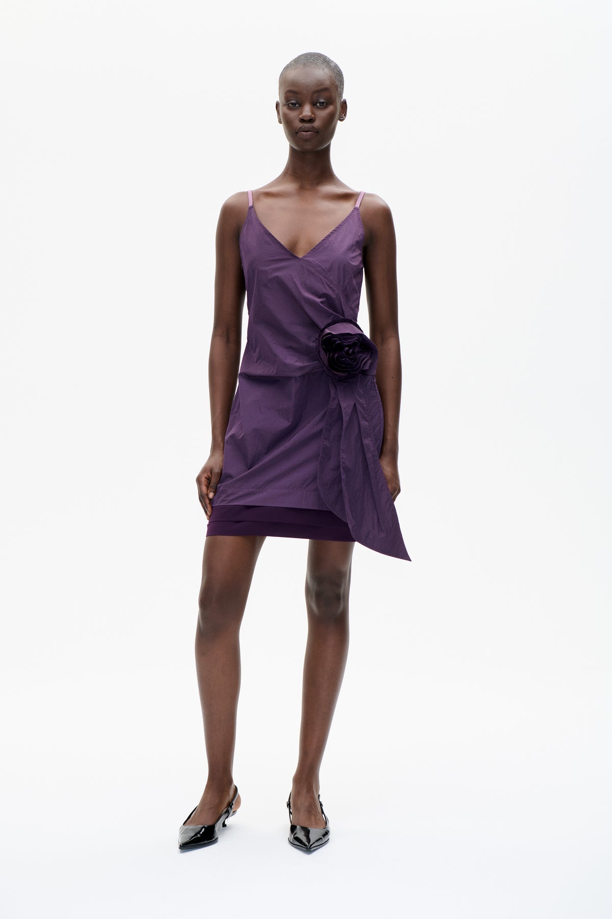 Front view of a model wearing a violet spaghetti-strap mini dress with a deep V neckline, sculptural rosette and draped hip panel.
