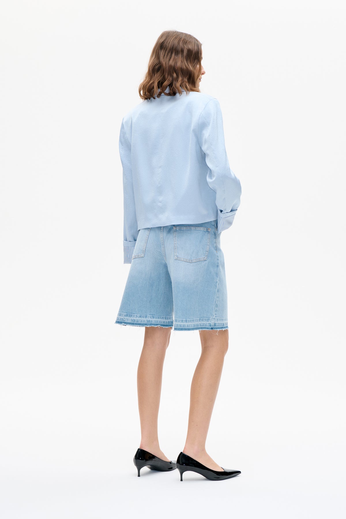 Rückenansicht eines Models, das eine hellblaue, strukturierte Langarmbluse mit gestreiften Bündchen und hellgewaschene denim-Shorts trägt.