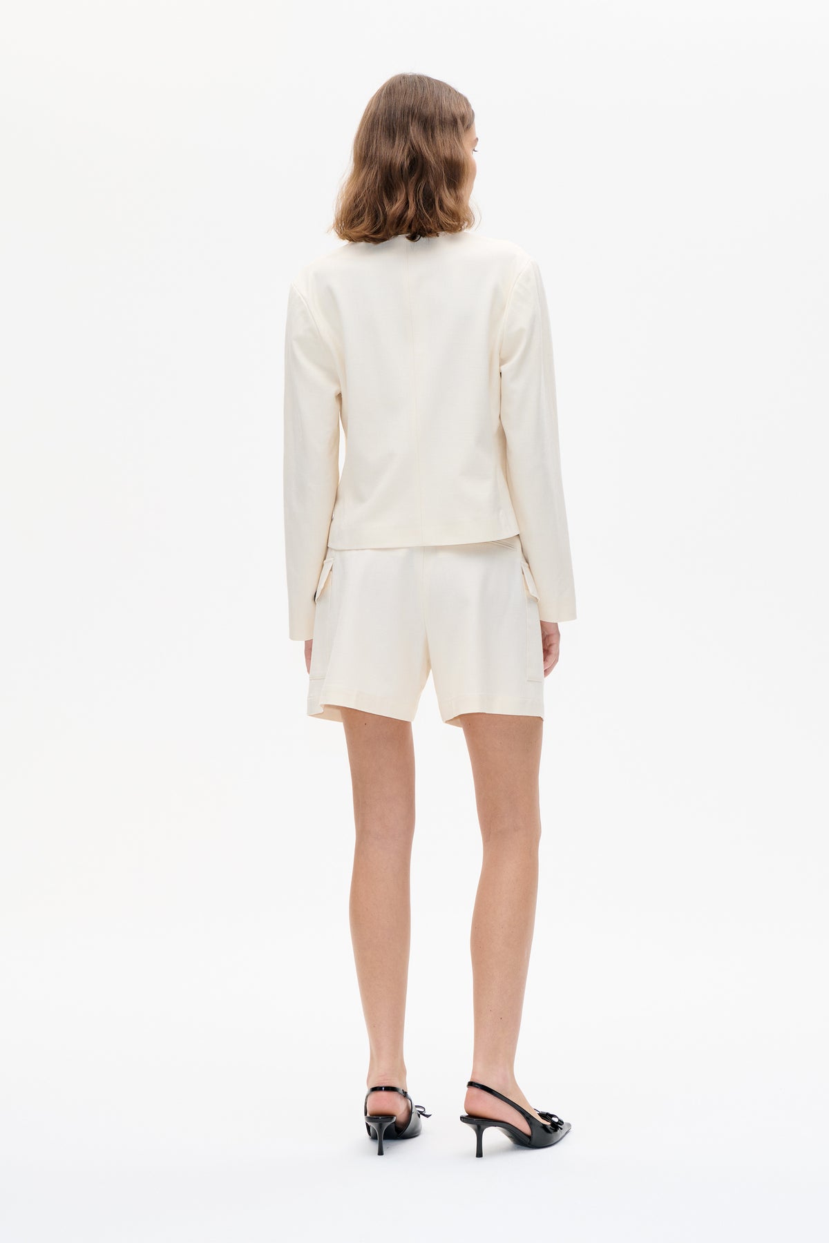 Rückenansicht eines Models in einer elfenbeinfarbenen, langärmeligen Jacke mit passenden, taillierten Shorts und schwarzen Slingback-Pumps mit Schleifendetail.