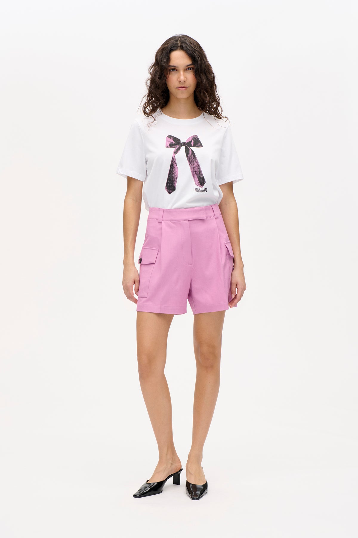 Front view of a white T-shirt with a pink-and-black bow graphic and small Baum und Pferdgarten logo, paired with pink cargo shorts.