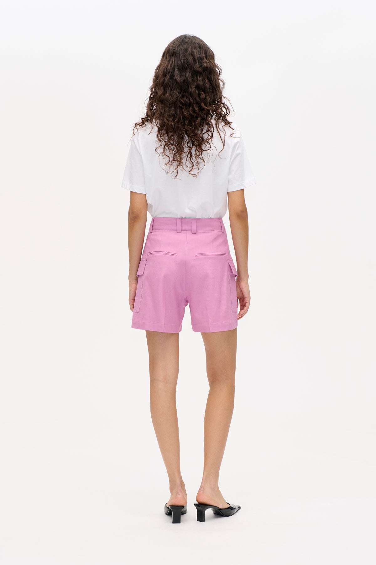 Back view of a model wearing a white short-sleeved T-shirt, pink high-waisted shorts with welt and flap pockets, and black heels.