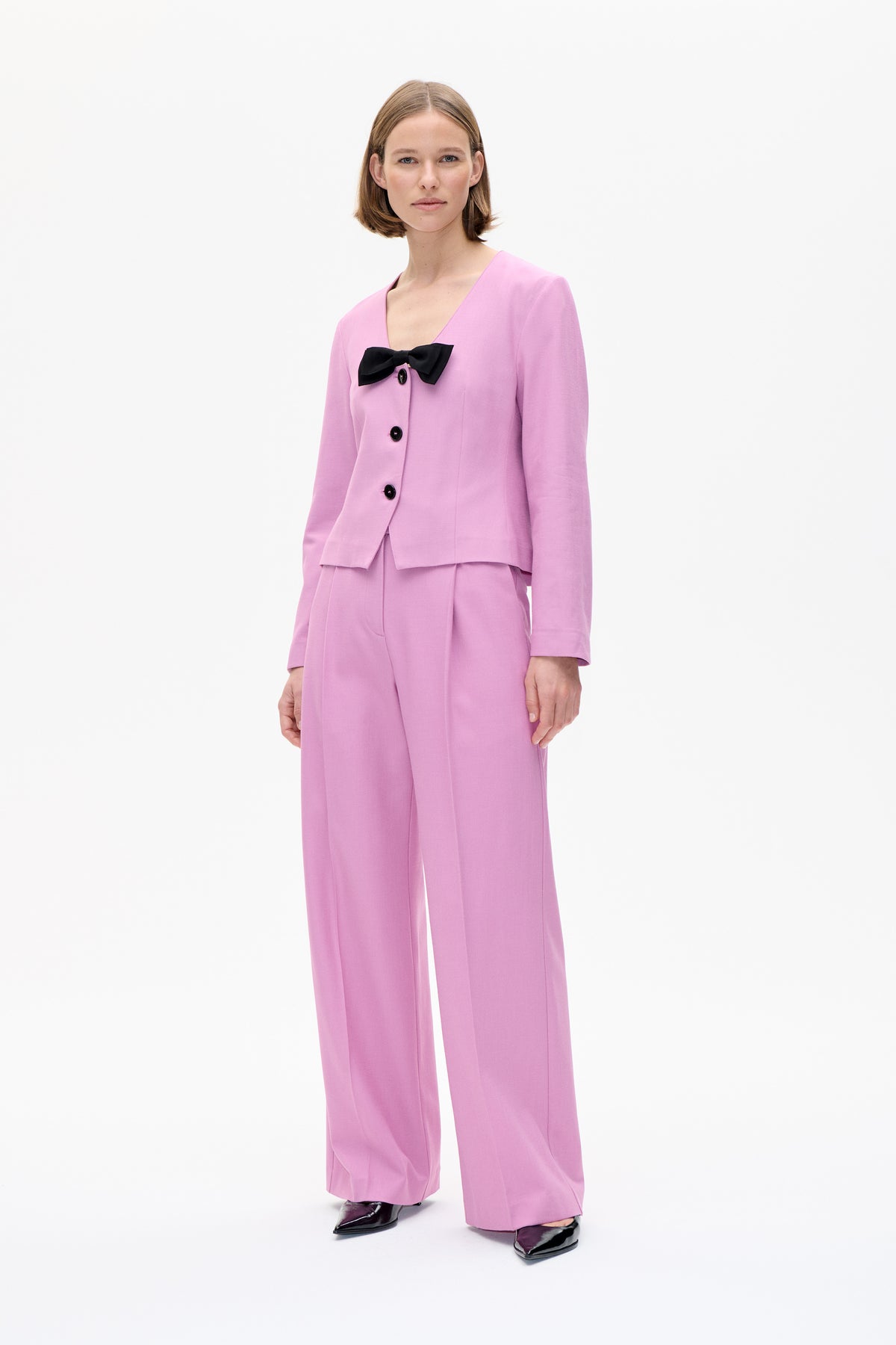 Model wearing a pink button-front jacket with a black bow detail and matching wide-leg trousers, shown from the front.
