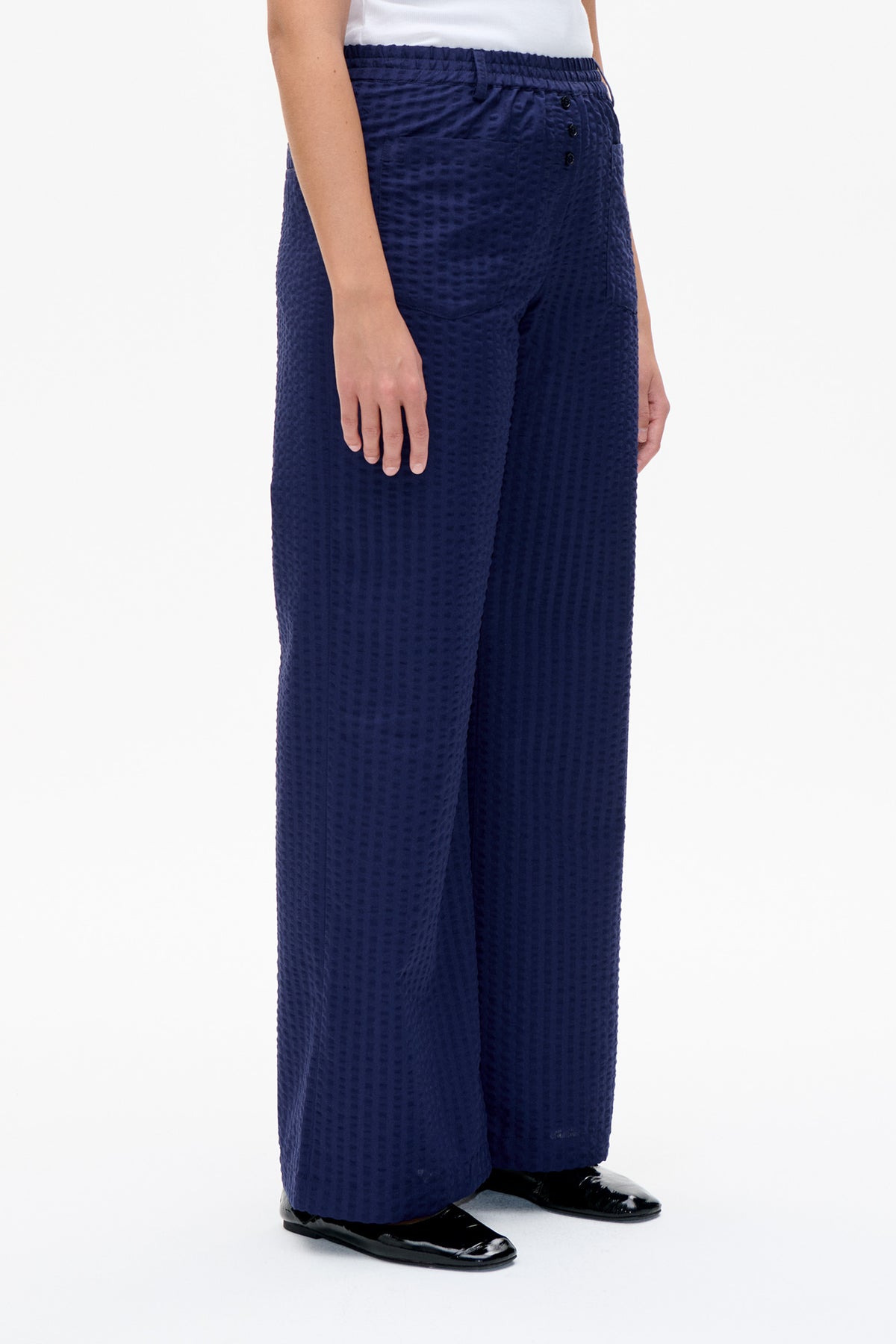 Close front view of navy wide-leg trousers with a raised textured pattern, gathered waistband and front buttons, worn with a white top.