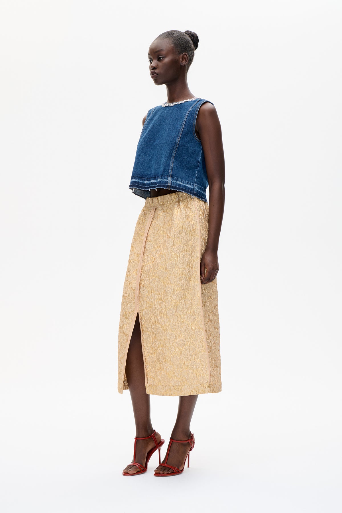 Model in a blue sleeveless denim top with embellished neckline and raw hem, gold textured midi skirt, and red heels against a white backdrop.