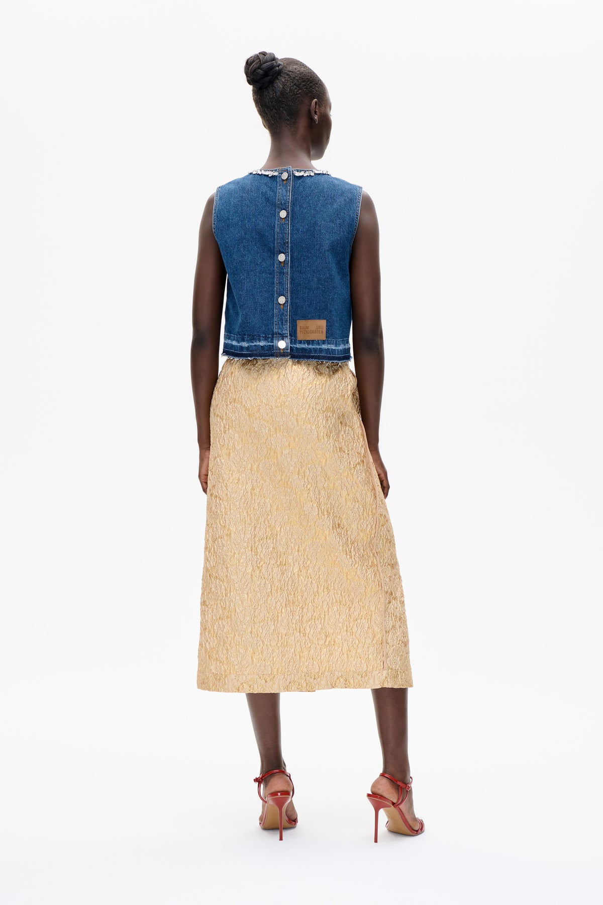 Rear view of a model wearing a blue sleeveless denim vest with a jeweled neckline and back buttons, paired with a pale gold textured skirt.