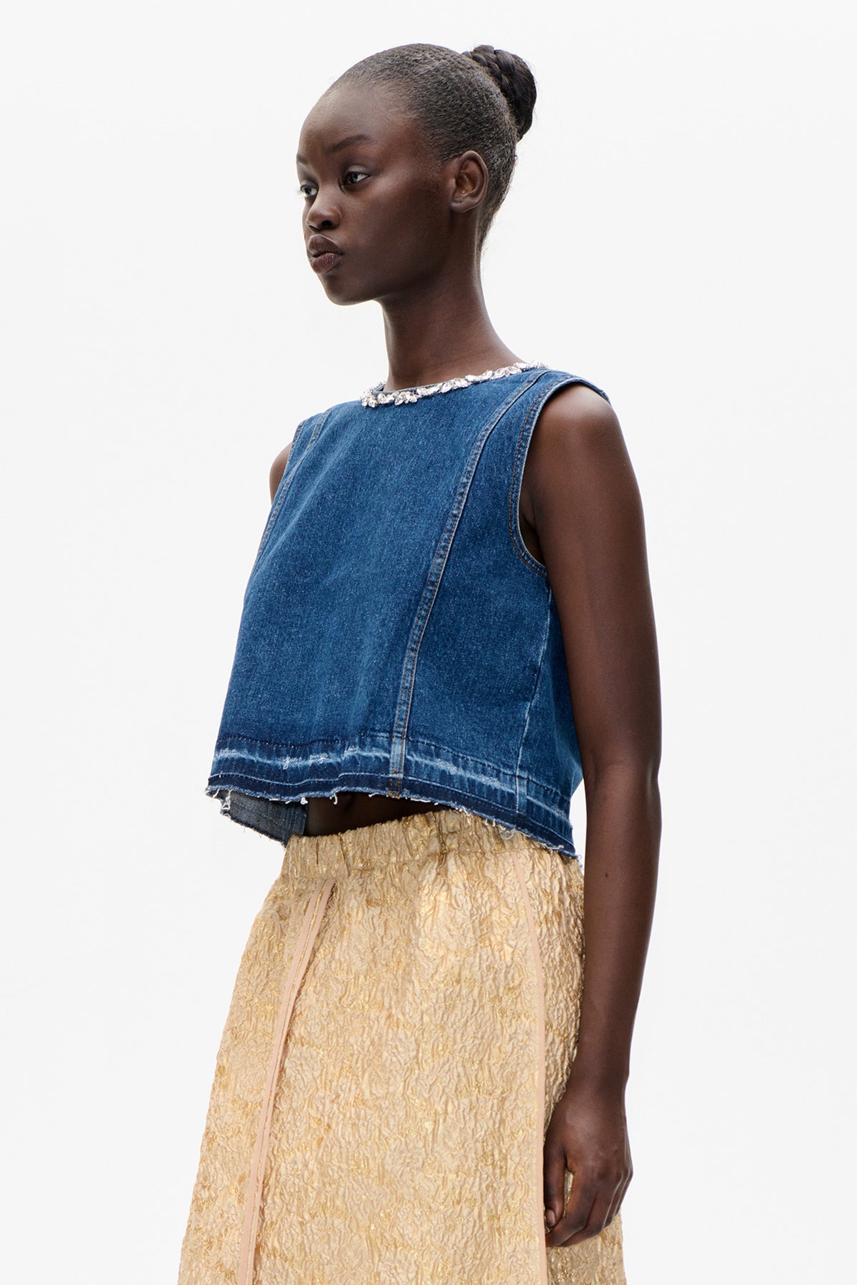 Model wearing a blue sleeveless denim top with a raw hem and jeweled neckline, paired with a textured gold skirt.