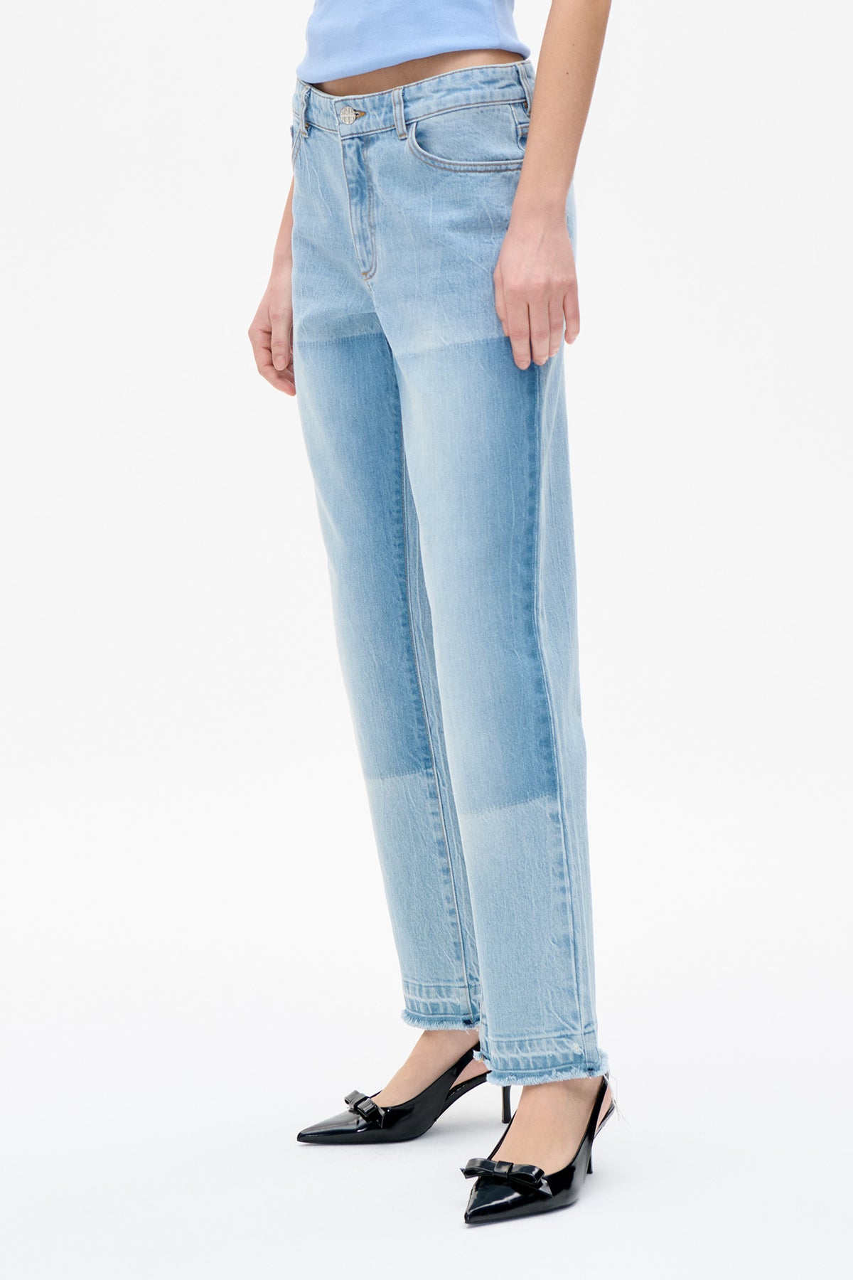 Close view of light-wash blue jeans with an asymmetrical button waistband, straight cropped legs and frayed hems, styled with black bow heels.