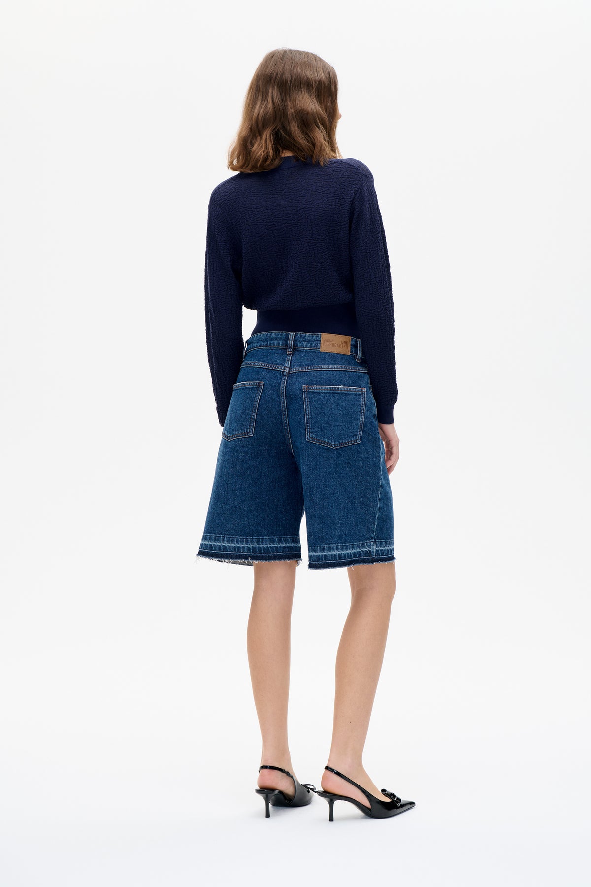 Achteraanzicht van een model in een marineblauwe, gestructureerde trui met lange mouwen en blauwe denim shorts tot op de knie met rafelige zomen.