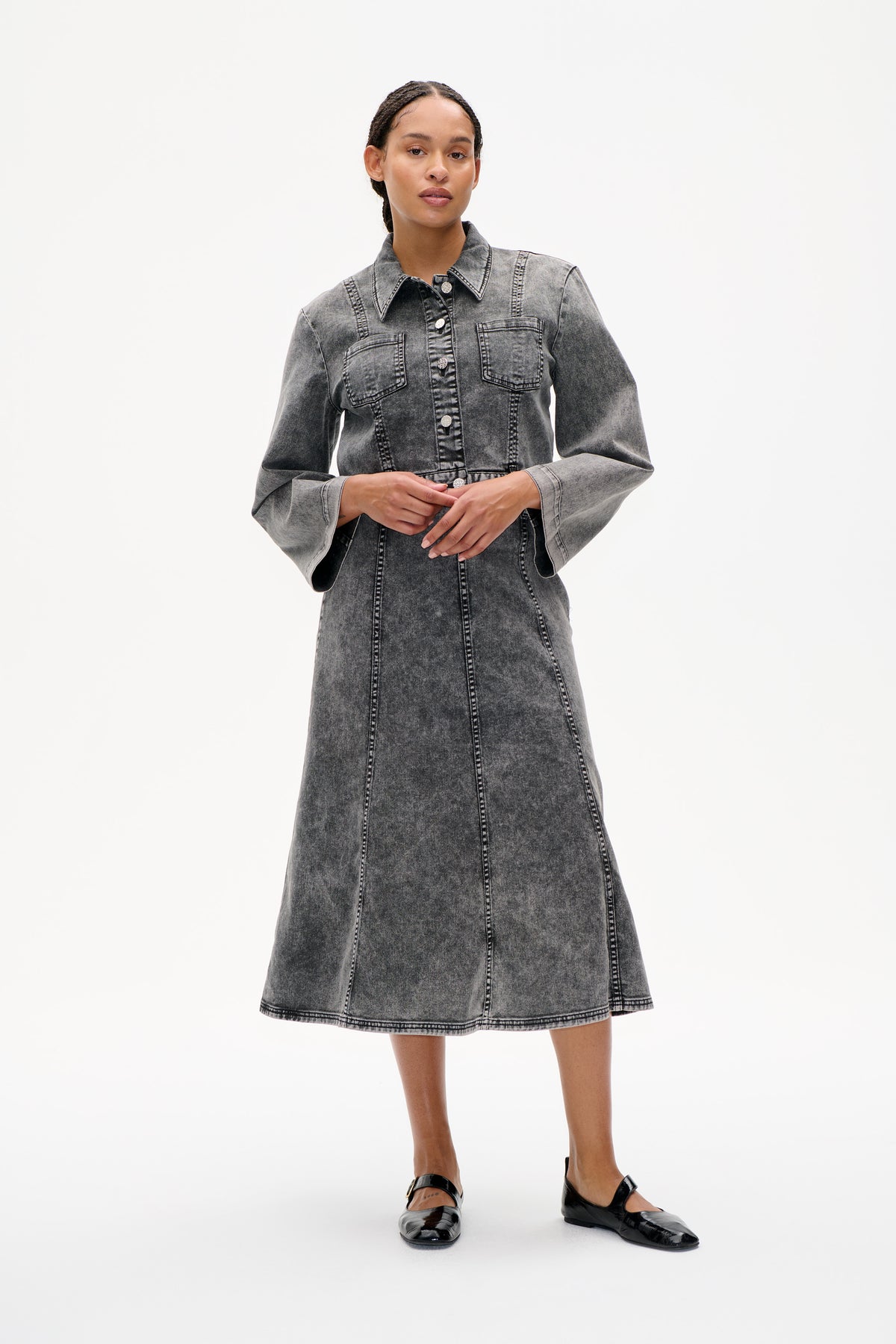 Model wearing a grey acid-wash denim shirt dress with a pointed collar, button front, chest pockets and contrast stitching, styled with black flats.