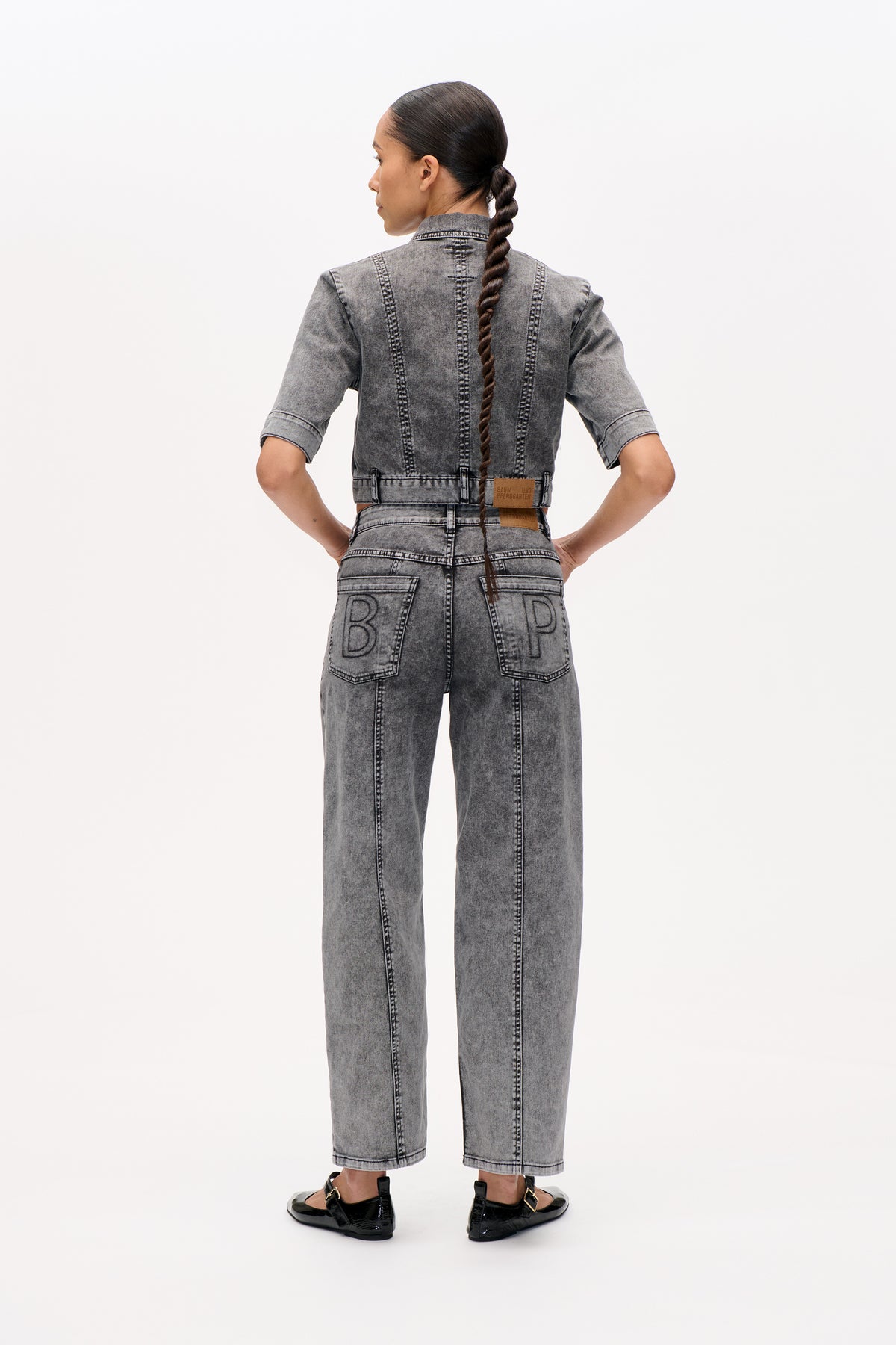 Back view of a model wearing a grey washed denim jacket with short sleeves and matching wide-leg jeans with outlined lettering on the pockets.