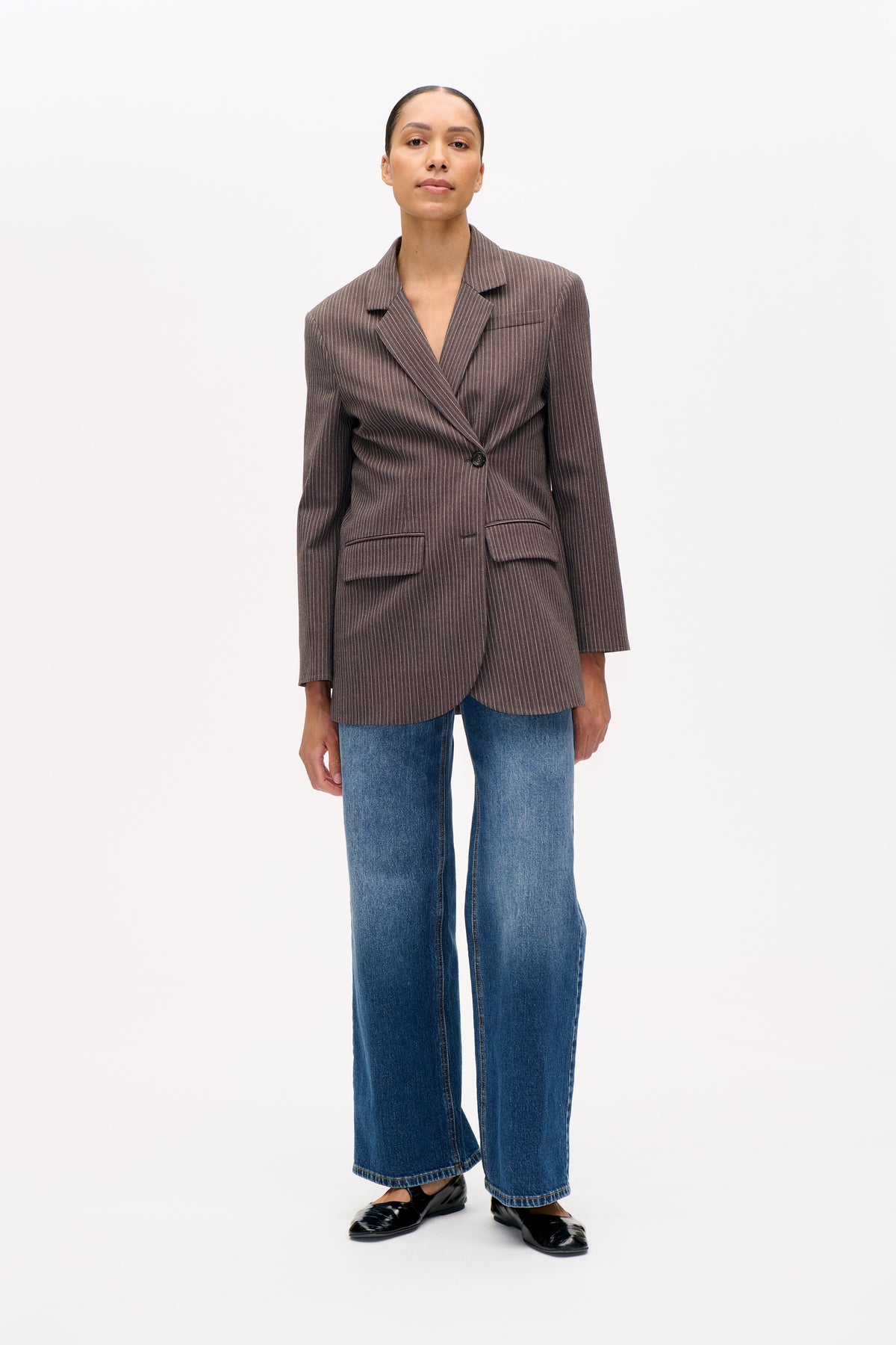 Model wearing a brown blazer with fine vertical stripes, notched lapels and flap pockets, paired with blue wide-leg jeans.
