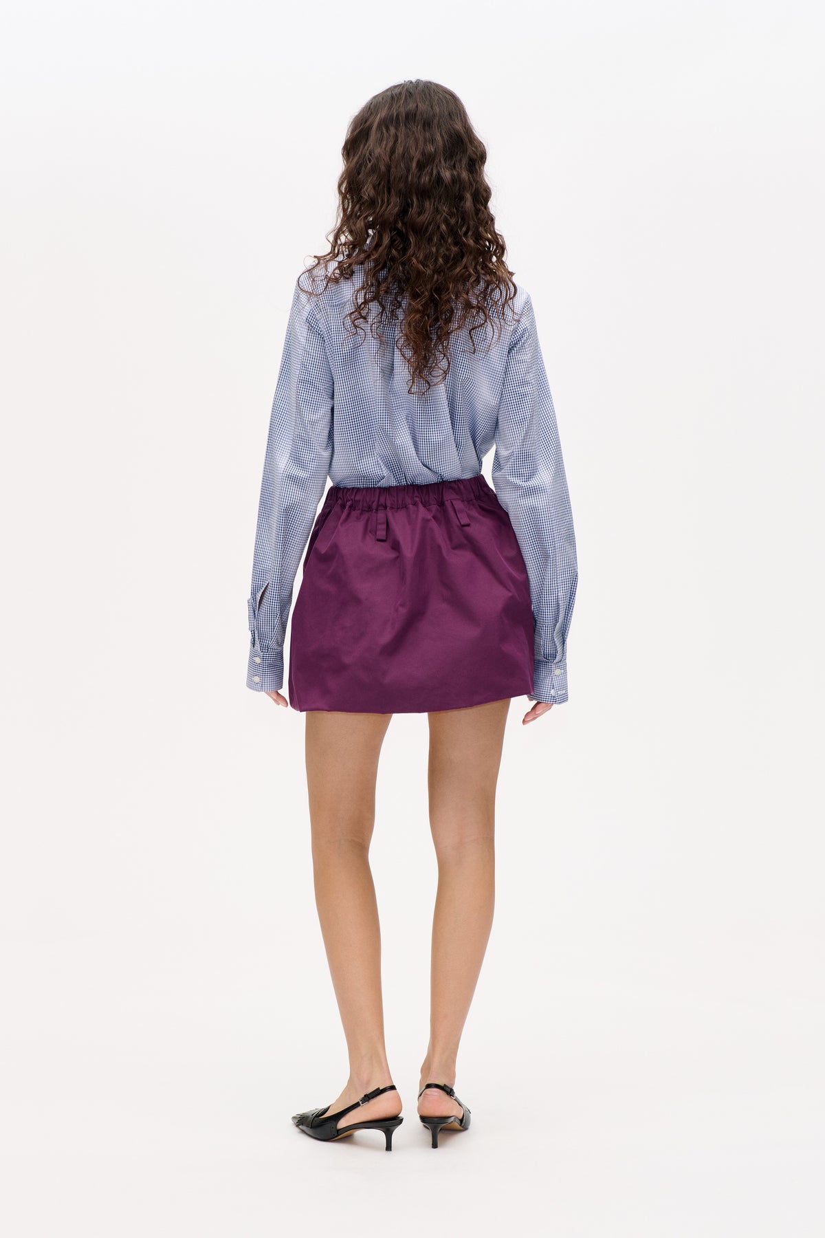 Rugaanzicht van een model dat een blauw-wit geruite blouse met lange mouwen en een pruimkleurige minirok met elastische taille draagt.