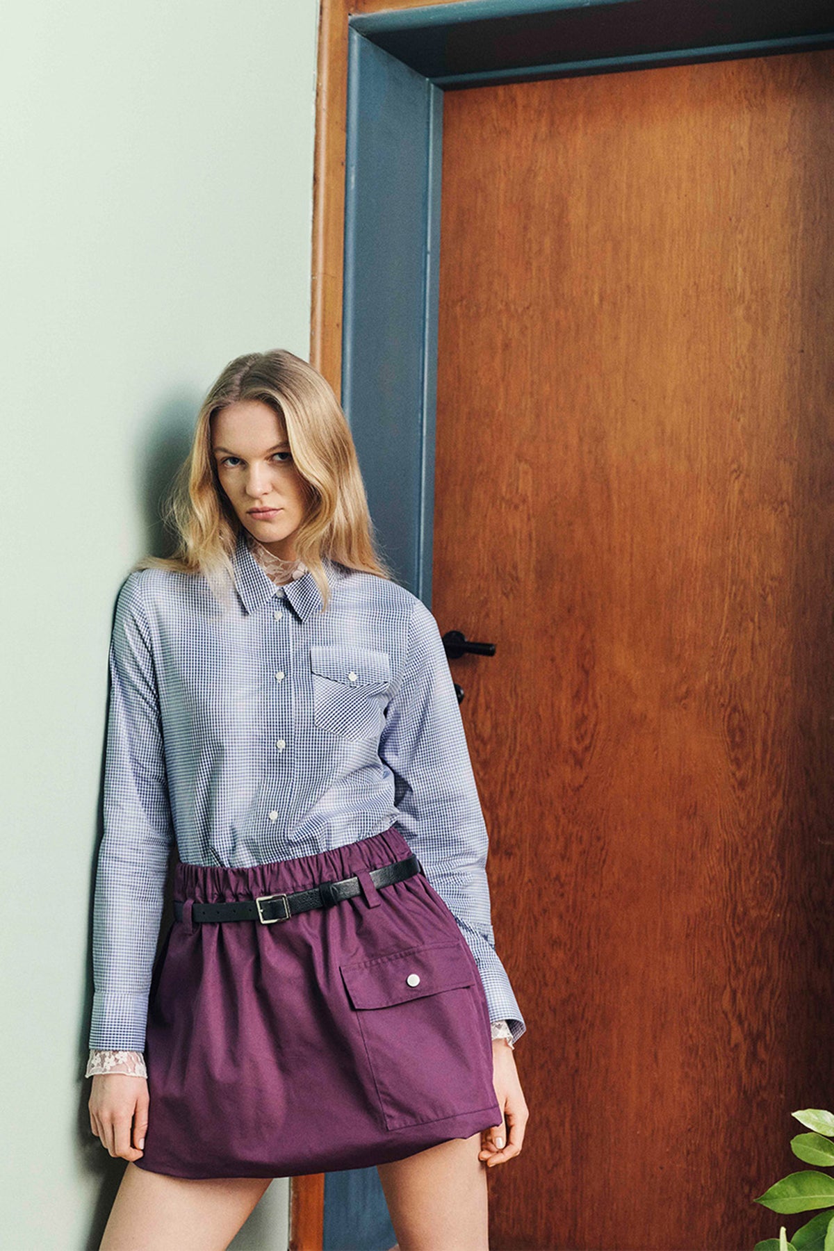 Model naast een houten deur draagt een blauw-wit geruite blouse met kant en een pruimkleurige utility-rok met ceintuur en klepzak