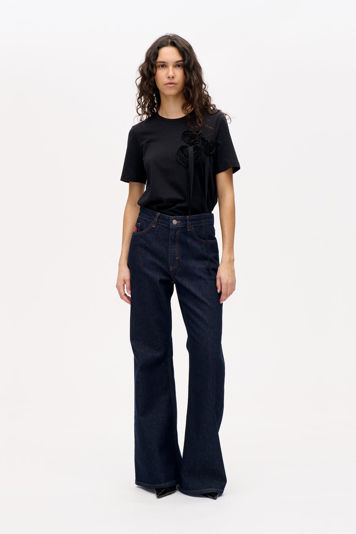 Front view of a model wearing a black short-sleeved T-shirt with layered fabric flower appliqués and dark wide-leg jeans.