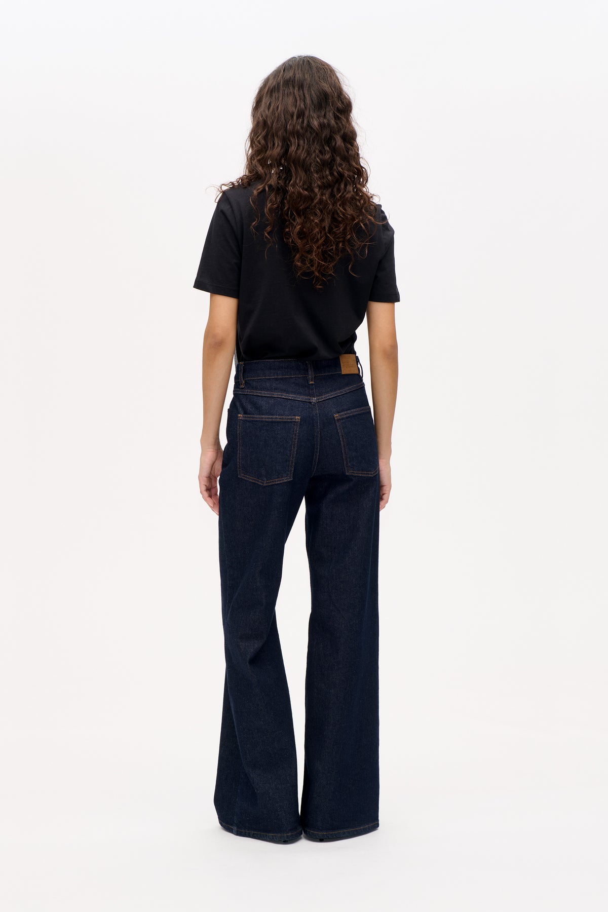 Rückenansicht eines Models, das ein schlichtes schwarzes kurzärmeliges T-Shirt und dunkelindigofarbene Jeans mit weitem Bein, kontrastierenden Nähten und Gesäßtaschen trägt.