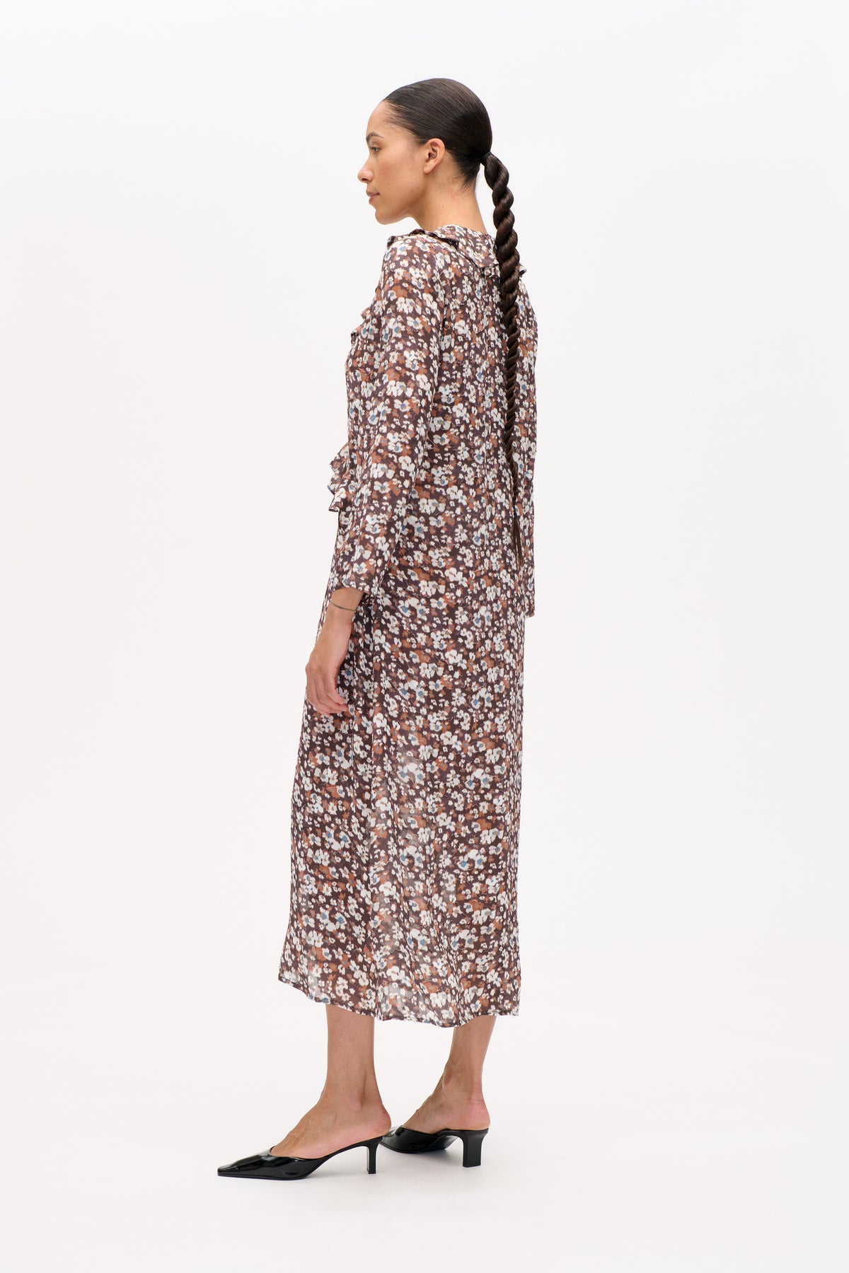 Side view of a model wearing a brown floral-print long-sleeved midi dress with ruffled trim and black heels.