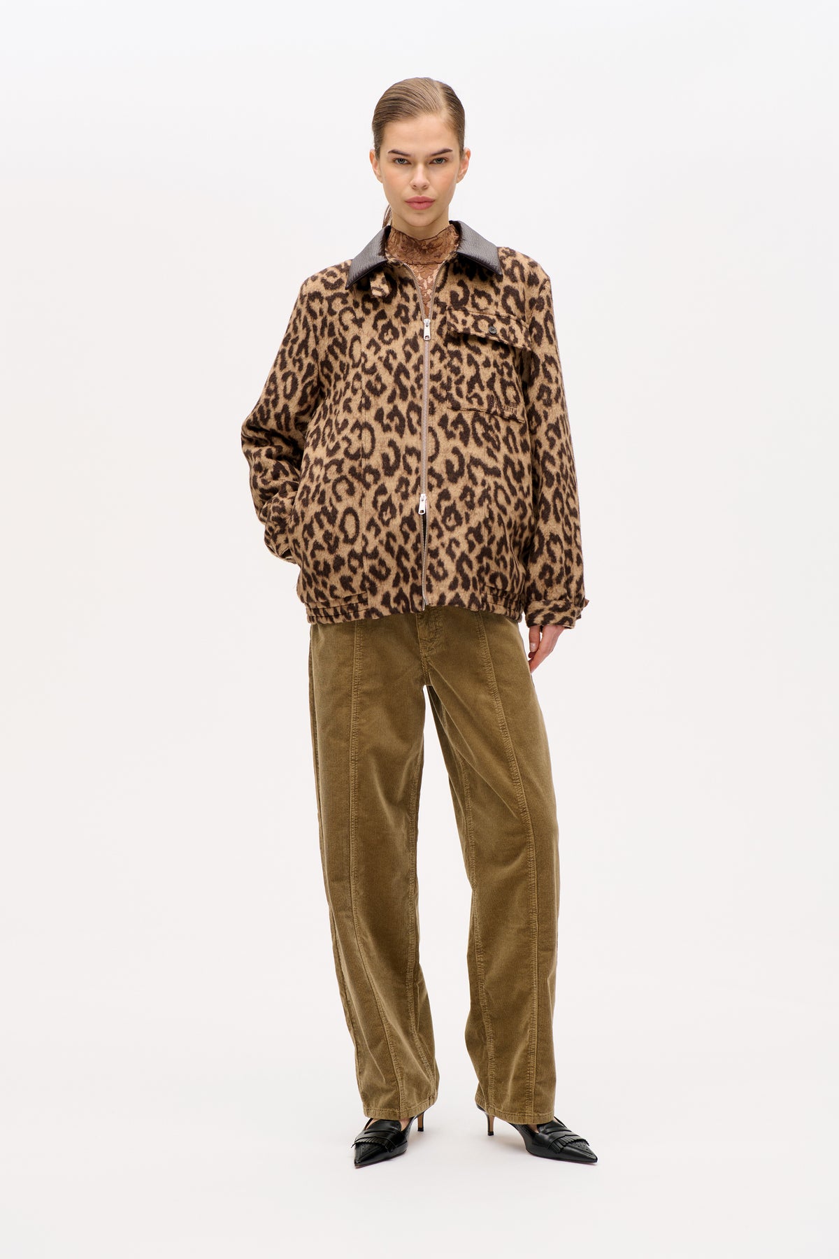 Model draagt een jasje met licht- en donkerbruine luipaardprint, rits aan de voorkant, zwarte kraag en olijfgroene ribfluwelen broek.