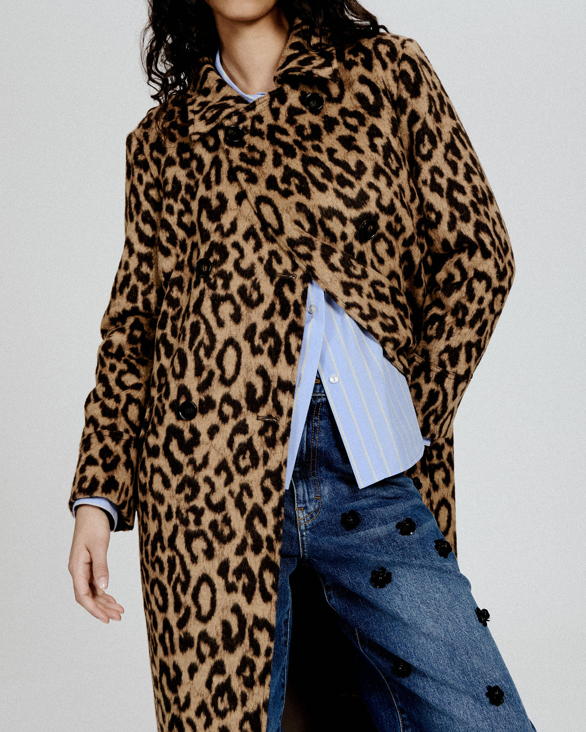 Cropped campaign portrait featuring a long brown and black leopard-print coat over a blue striped shirt and embellished jeans.